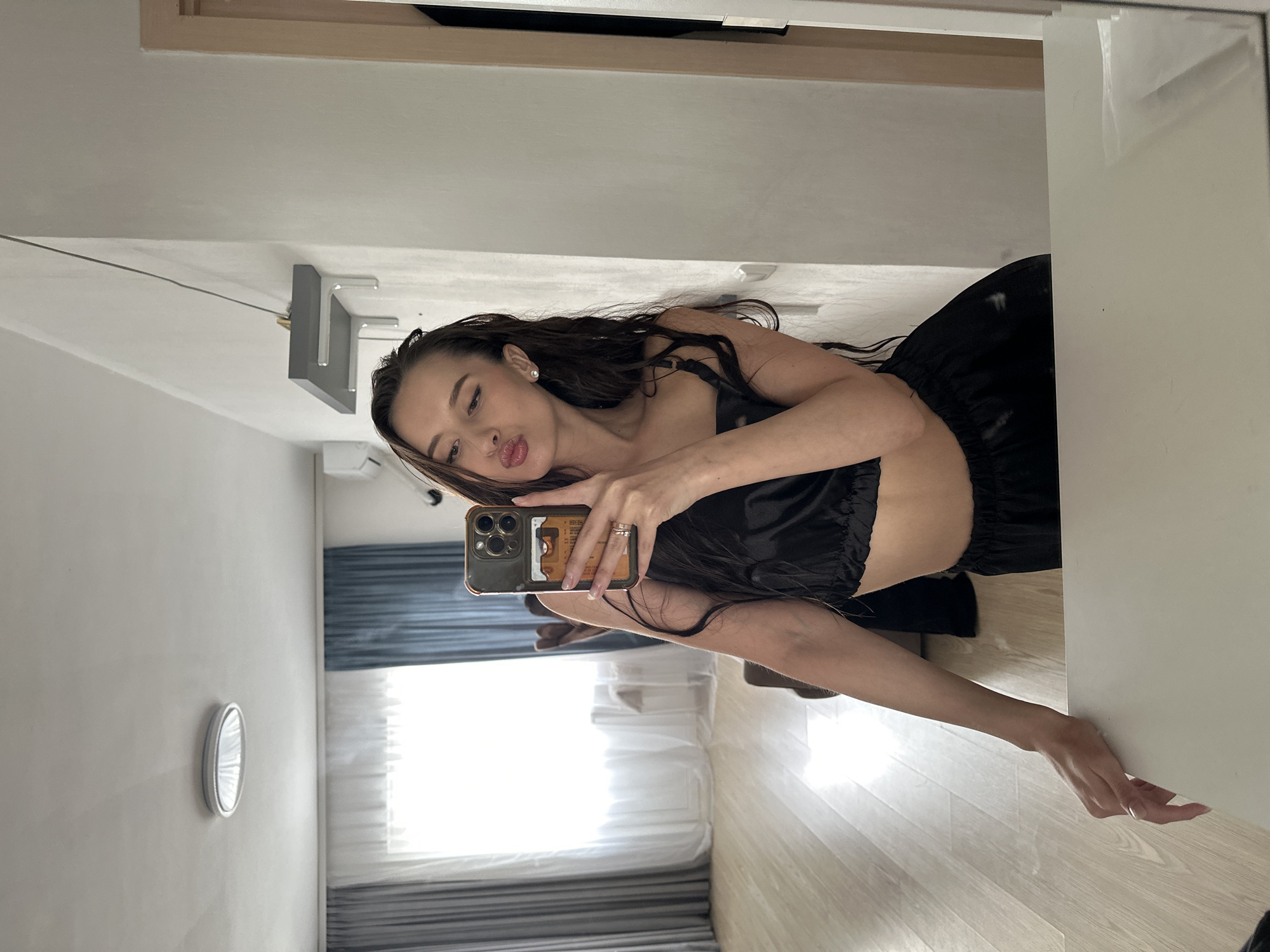 A woman is taking a selfie in a room. She is wearing a black top and skirt. A mirror is to her right. The room has a window with blinds. The location is likely inside a residential building. There is no information to indicate that the location is in the city of Yekaterinburg, Russia.
