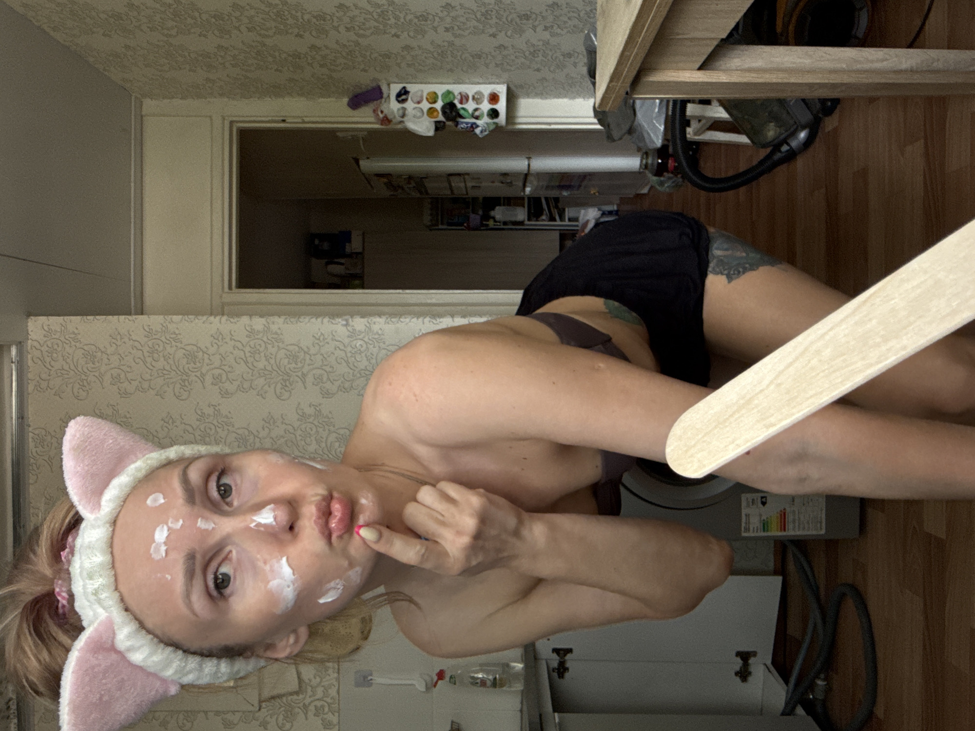 A woman with a face cream and pink cat ears is in a room. She has a finger on her lips. The room appears to be a kitchen, with a sink, cupboards and a refrigerator visible. The image was taken in a domestic setting. The location may be in Chaikovsky, Russia.