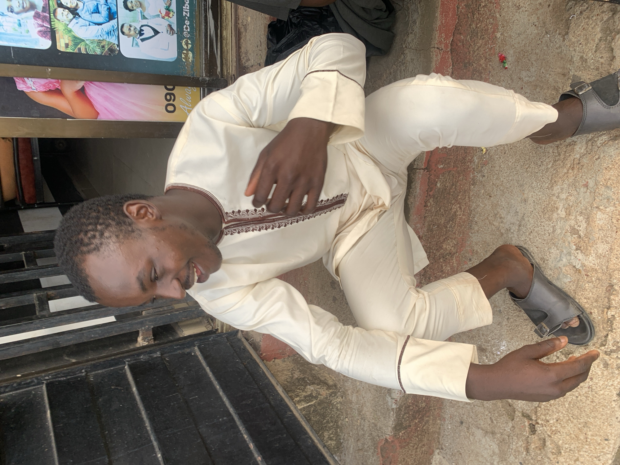 A man is kneeling on a concrete surface outside a shop in Barakin Akawo, Nigeria. He is wearing a light-colored, long-sleeved tunic and pants, and grey sandals. He is turned to the left, smiling and looking downward. He is positioned next to a black metal grating, and near a shop front which has posters of people in formal wear.