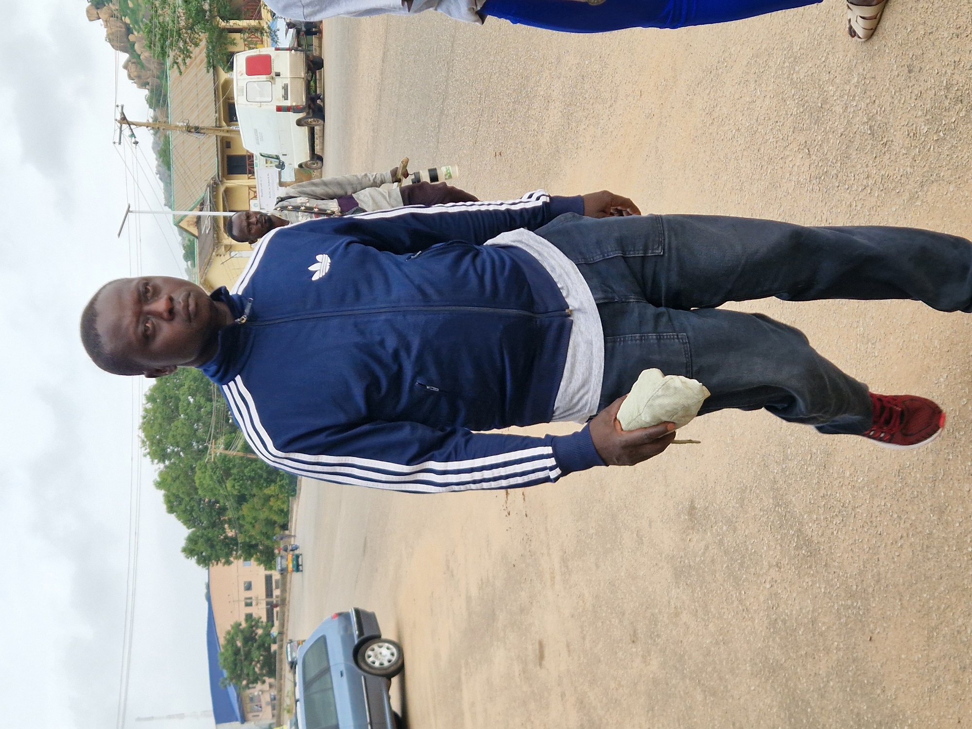 The image shows a man in the foreground walking on a dusty road. He is wearing a dark blue tracksuit with white stripes on the sleeves and a light-colored t-shirt underneath. He is also wearing dark blue jeans and red sneakers. The man is holding a light-colored cloth in his right hand. In the background, there are several buildings, trees, and vehicles, including a white van and a blue station wagon. The sky is overcast. The image was taken in Kabong, Nigeria.