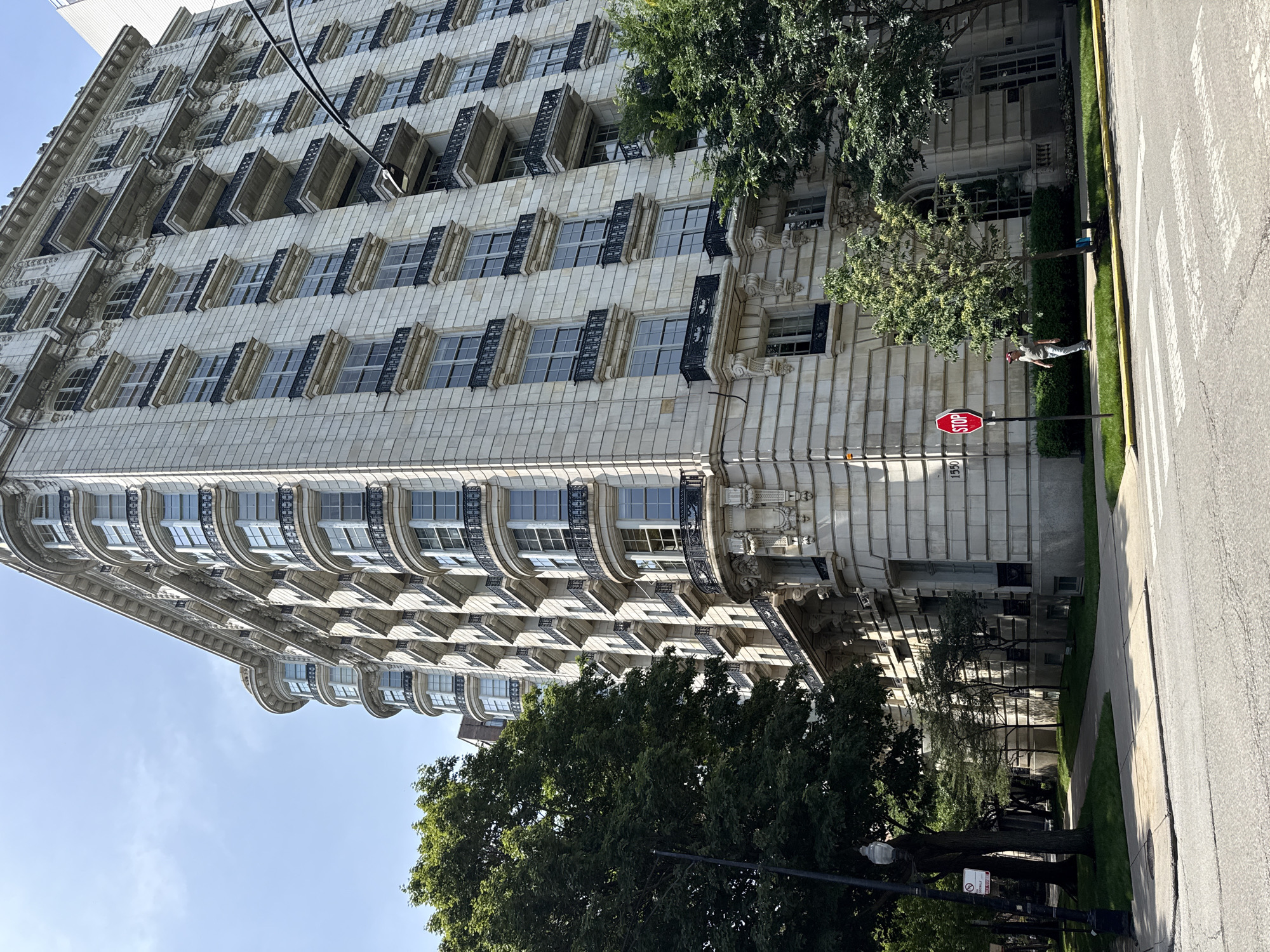 The image shows a low-angle, full-shot view of the historic apartment building at 1550 N. Sandburg Terrace in Chicago, Illinois, United States of America. The building, constructed of light-colored stone with ornate architectural details, rises several stories high, featuring numerous windows and balconies with black wrought-iron railings. The facade is characterized by a series of bay windows and decorative carvings above some of the windows and balconies.
In the foreground, a paved street with a crosswalk is visible, indicating a pedestrian crossing area. To the left of the street, a sidewalk lines a grassy area with trees. A street lamp with an ornate top stands tall, and a "No Parking" sign is attached to its pole. A red octagonal "STOP" sign is positioned on a pole near the corner of the building.
A person, dressed in a light-colored shirt, dark pants, and a cap, is walking on the sidewalk near the stop sign, facing away from the camera. Lush green trees with full foliage partially obscure the lower floors of the building, framing the structure and adding to the urban park-like setting. The sky above is clear with a few wispy clouds. The overall scene depicts a sunny day in a well-maintained urban environment, highlighting the grandeur of the building and the presence of urban greenery.