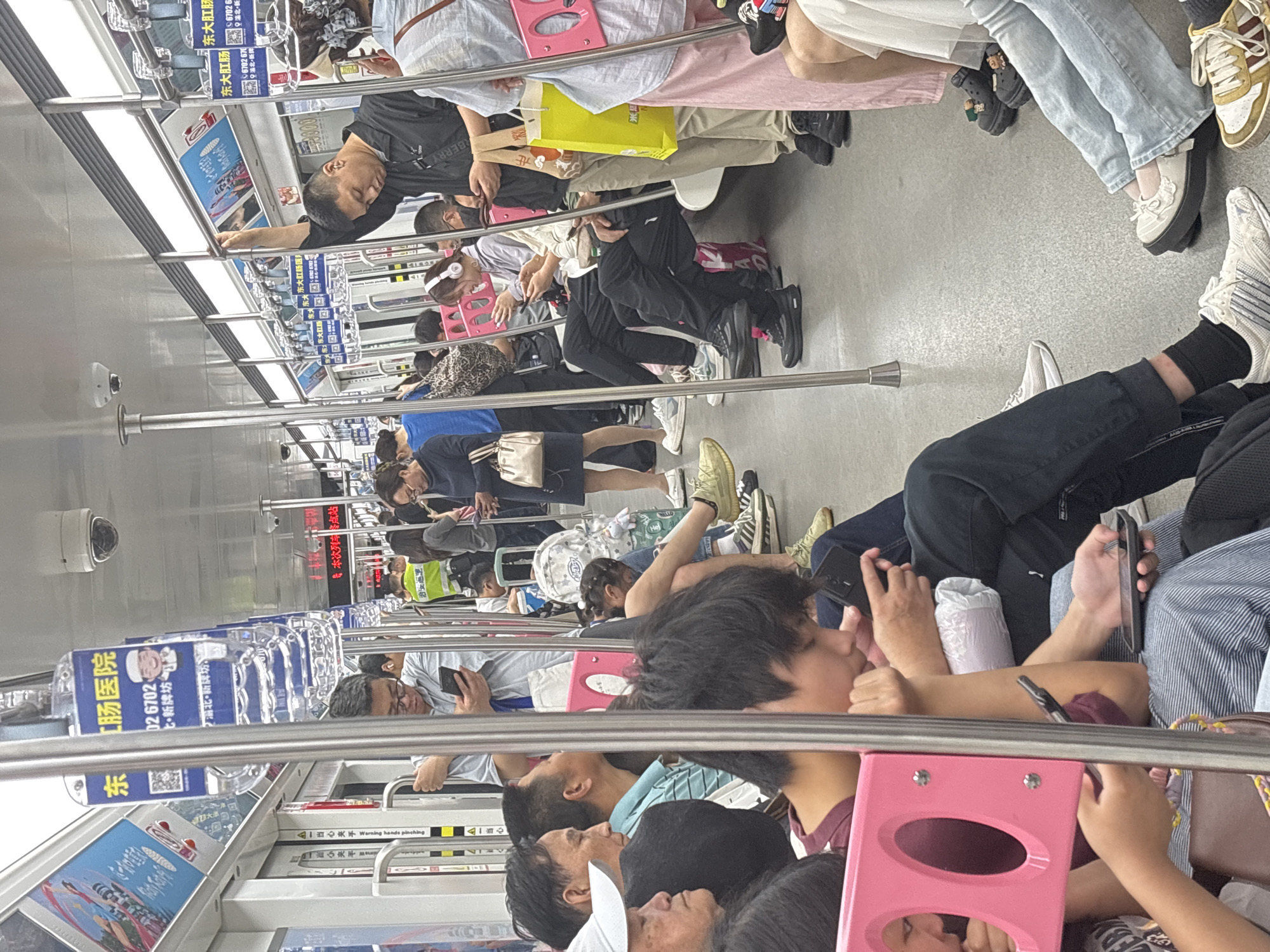 The image shows a crowded subway car in 龙溪街道, China. Passengers are sitting and standing, with many using their cell phones. The interior of the subway car has advertisements and safety warnings posted. Several passengers are leaning on the handrail.