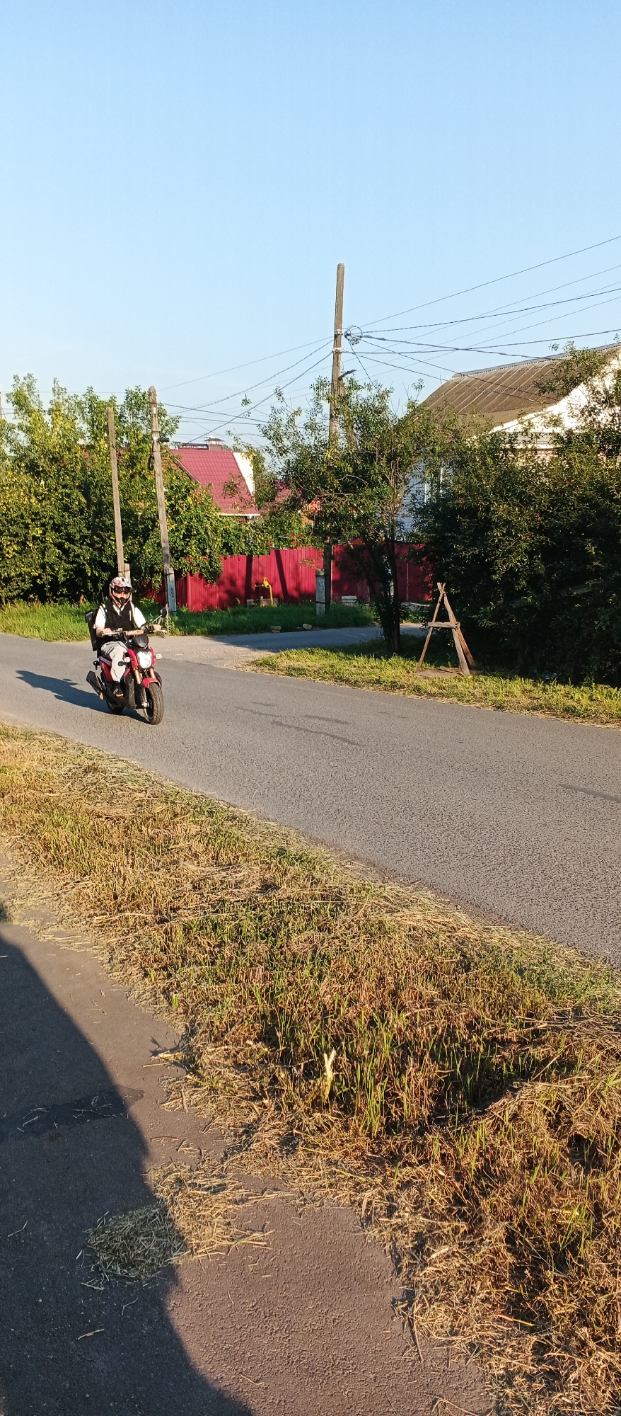 A person rides a red and white motorcycle on a paved road, heading towards the viewer. The rider is wearing a helmet, a dark vest, and light-colored pants. The motorcycle has a white windshield and a black seat. The road is bordered by a grassy area on the left and a more overgrown, dry grassy area on the right. In the background, there are several houses with red roofs and white siding, a red fence, and trees. Utility poles with numerous wires are also visible. The sky is clear and blue. The lighting suggests it is daytime, likely late afternoon due to the long shadows. This scene is consistent with a residential area in Izhevsk, Russia.
