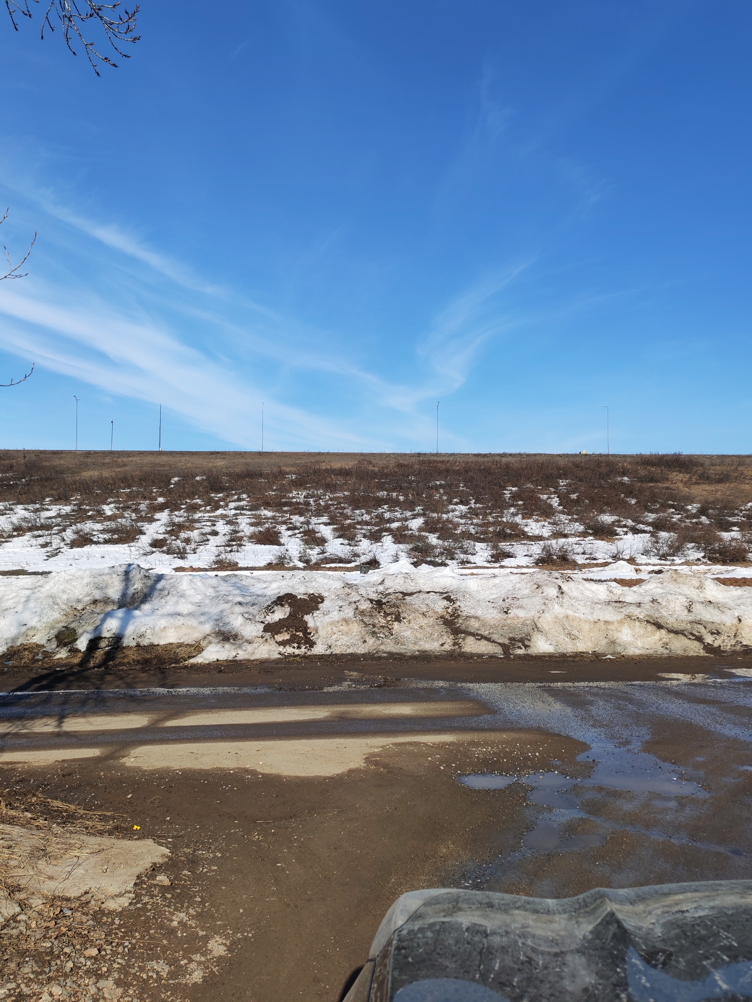 The image depicts a partly snow-covered landscape under a clear blue sky with thin, streaky clouds. In the midground, there is a raised embankment covered with dry vegetation and patches of melting snow. Streetlights are visible along the top of the embankment. The foreground shows a muddy area with puddles and remnants of snow and debris. A portion of a vehicle's tire is visible in the lower right corner. The specific location in Chaikovsky, Russia is undeterminable from the visual content of this image.