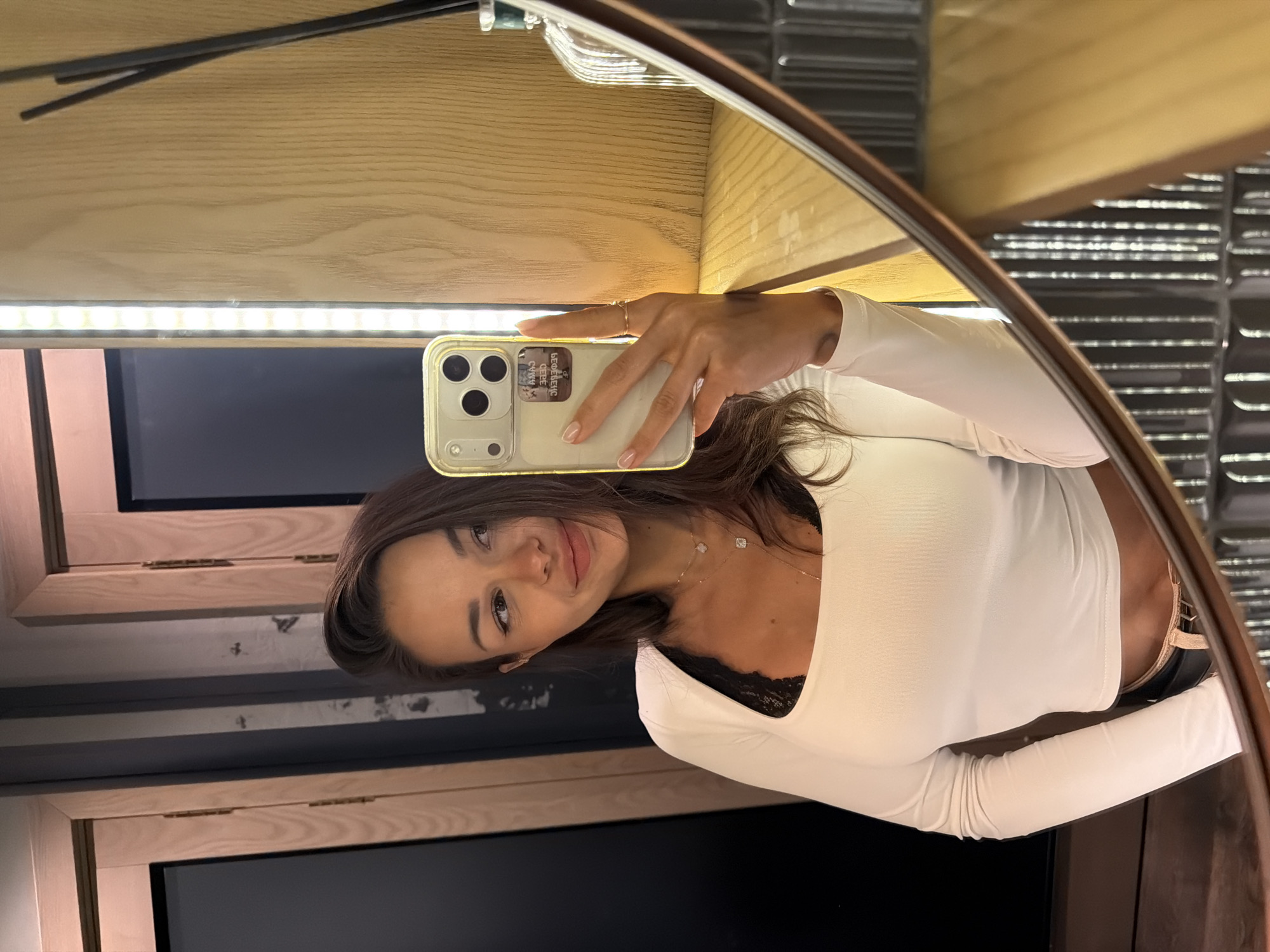 A young adult female, appearing to be of Eastern European descent, is taking a mirror selfie in what appears to be a public restroom or fitting room. She is wearing a white long-sleeved crop top with black lace trim, black pants, and a thin belt. She has long, dark brown hair and is wearing light makeup, including eyeliner and lip gloss. She is holding a smartphone with a light-colored case in her right hand, angled to capture her reflection. She is wearing two delicate necklaces, one with a small square pendant and another with a small spherical pendant. She has a thin gold ring on her left ring finger. The background consists of a wooden paneling, a mirrored surface reflecting the subject, a strip of LED lighting, and what appears to be a diffuser with reeds on a shelf. The overall setting suggests a modern, clean aesthetic. The image does not provide specific visual cues to definitively place it in Saint Petersburg, Russia, or any other specific city.