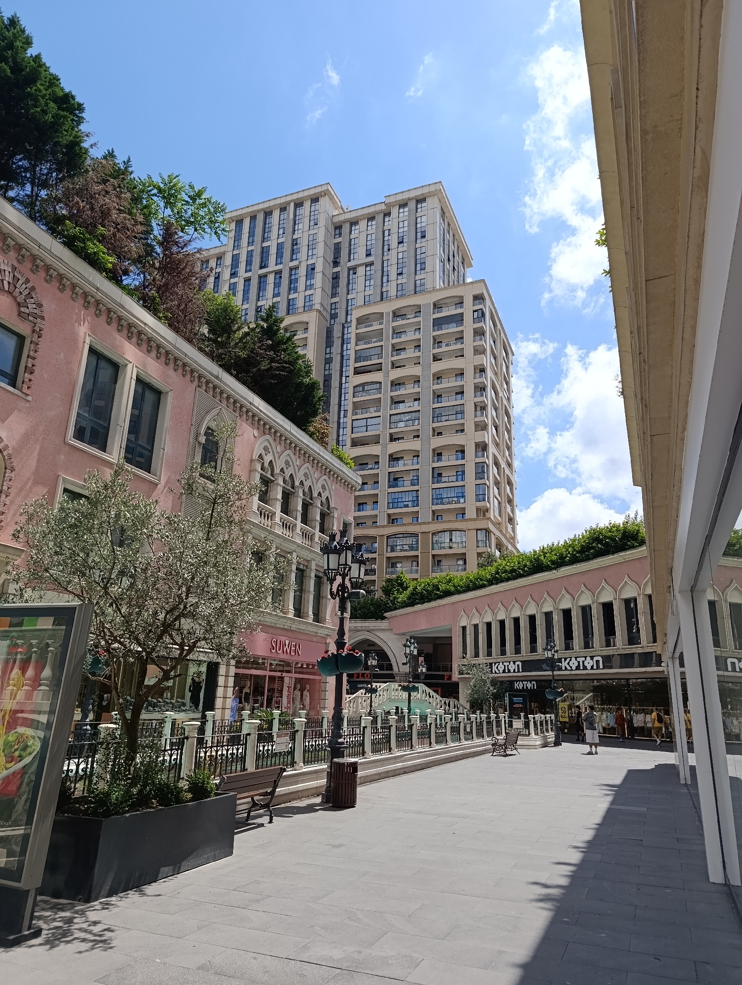The image depicts a modern shopping street in Gaziosmanpaşa, Turkey, under a clear blue sky with scattered white clouds. In the foreground, a paved walkway is lined with shops and architectural elements. A pink building with arched windows and decorative white trim is visible on the left. A small tree with delicate foliage is planted in a dark planter box. A park bench and a lamppost are also present. In the background, two tall, modern apartment buildings with numerous balconies dominate the skyline. Shops with glass storefronts are visible, including one with the "KOTON" logo. Several individuals are walking on the street, and some are entering or exiting shops. The overall scene suggests a vibrant urban environment with a mix of traditional and contemporary architectural styles.