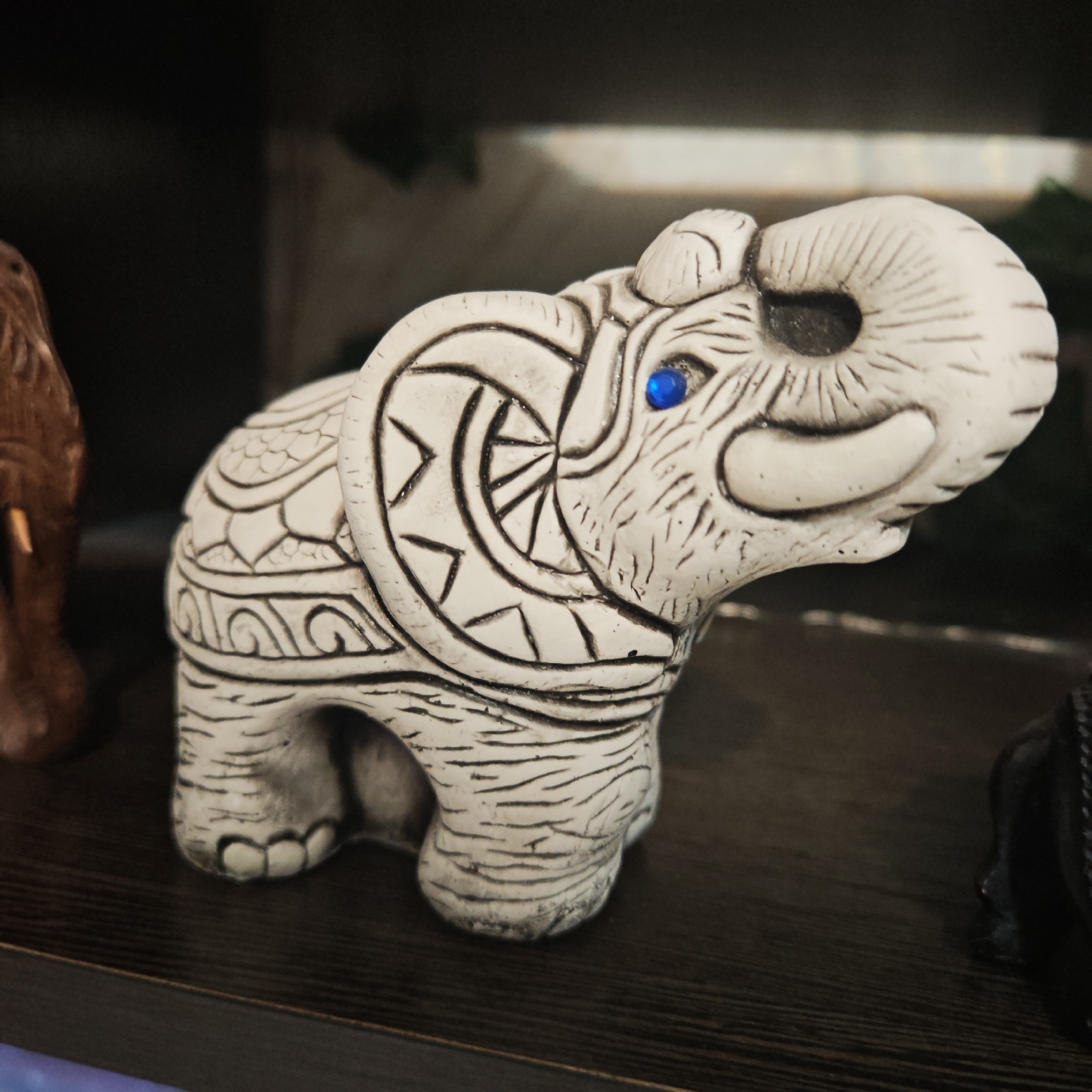 A close-up shot depicts a small, ornate elephant figurine with a blue gemstone eye, resting on a dark wooden surface. The figurine is primarily white or off-white with detailed etched designs in black. The location or the city of Rostov, Russia is not evident in the image. The focus is entirely on the figurine and the immediate surface it occupies. Other vague objects are partially visible on either side of the elephant, but they are out of focus and difficult to identify. The background is dark.
