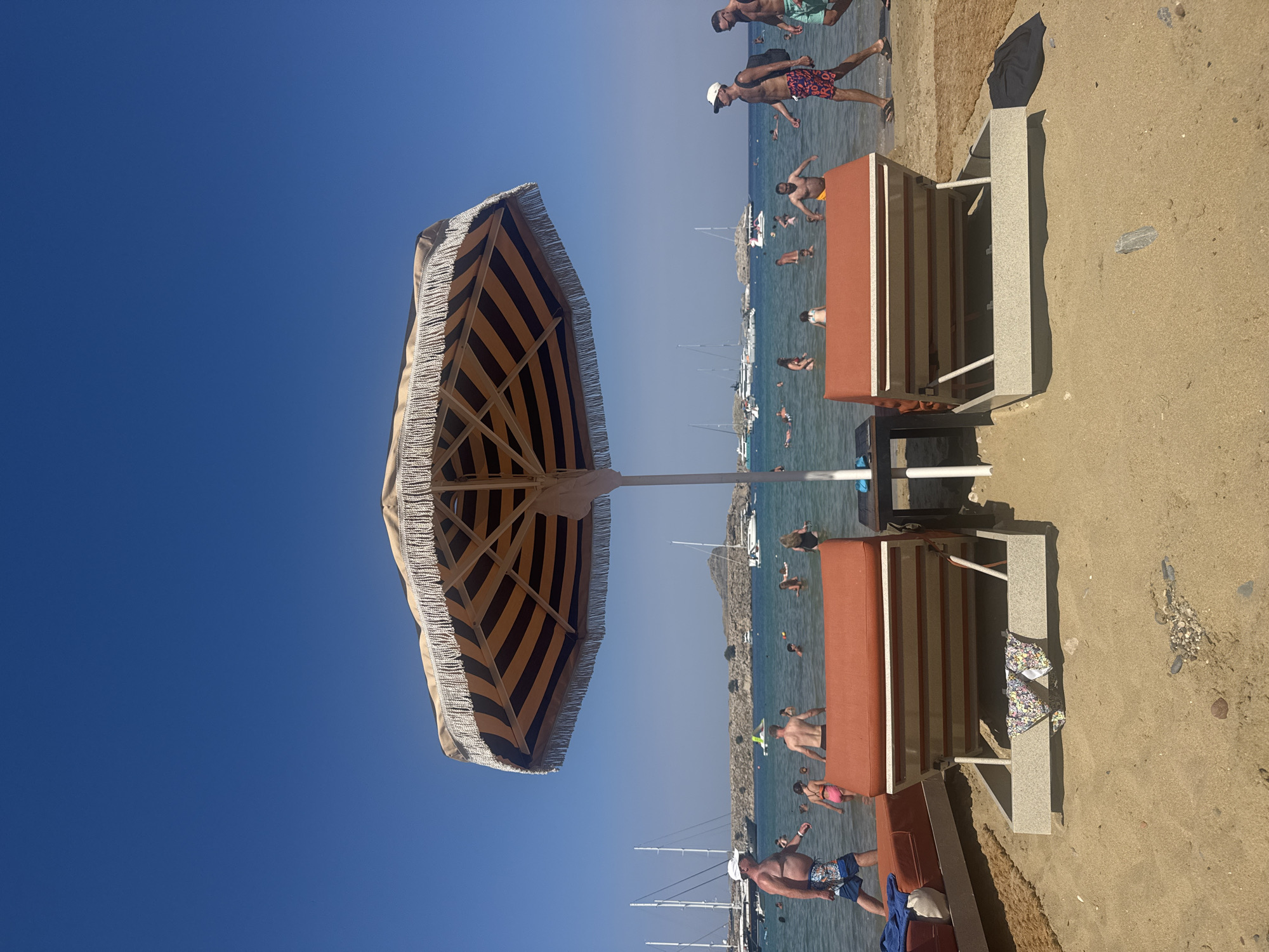 The image depicts a sunny day at a beach in Lindos, Greece. In the foreground, two orange lounge chairs are set up on the sand, partially covered by a large, striped umbrella. A small table is situated between the chairs. Beachgoers are visible in the shallow, clear water, with several boats and yachts anchored in the bay. The sky is a vibrant, clear blue. The overall scene suggests a relaxed atmosphere at a popular tourist destination.