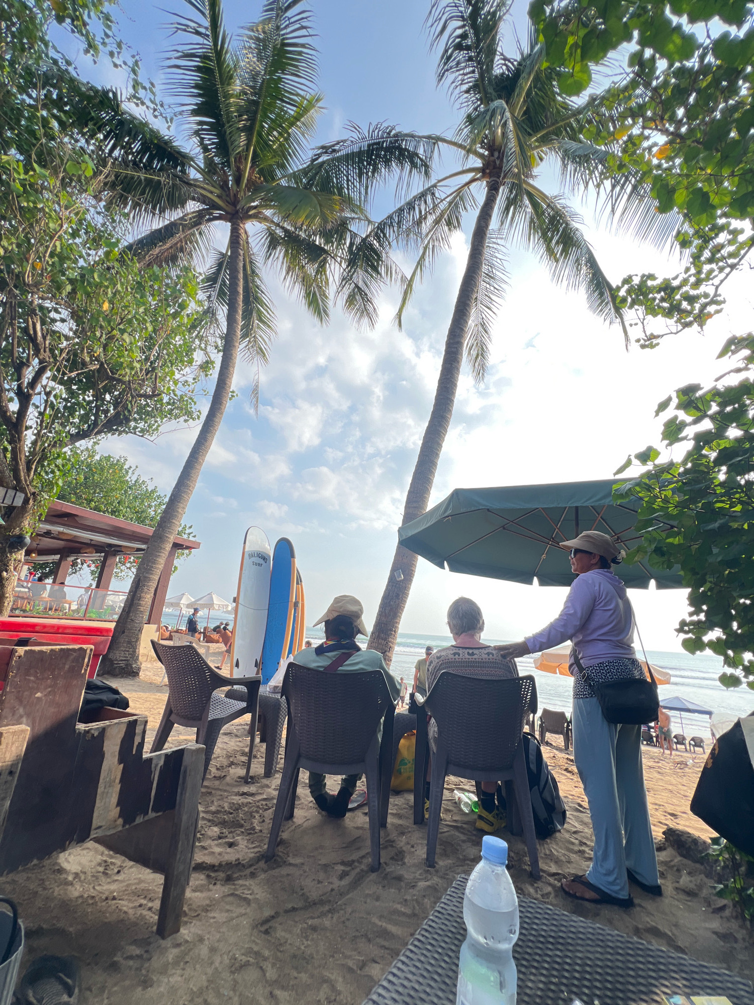 The image depicts a beachfront scene in Kuta, Indonesia, under a partly cloudy sky. In the foreground, a plastic water bottle sits on a table made of woven material. Two people are seated on chairs in the middle ground, facing away from the camera. One individual, wearing a light purple hoodie, light blue pants, and sandals, stands and places a hand on the shoulder of the seated person, who is wearing a patterned sweater. A large green umbrella is positioned behind them, providing shade. Two surfboards stand upright on the sand between the seated individuals and the ocean. In the background, a beach bar with red siding and a brown roof is visible, with people seated at tables. Further in the distance, beach umbrellas and loungers are scattered along the sandy shore, with the ocean in the far background. Palm trees are prominent throughout the image, framing the scene.