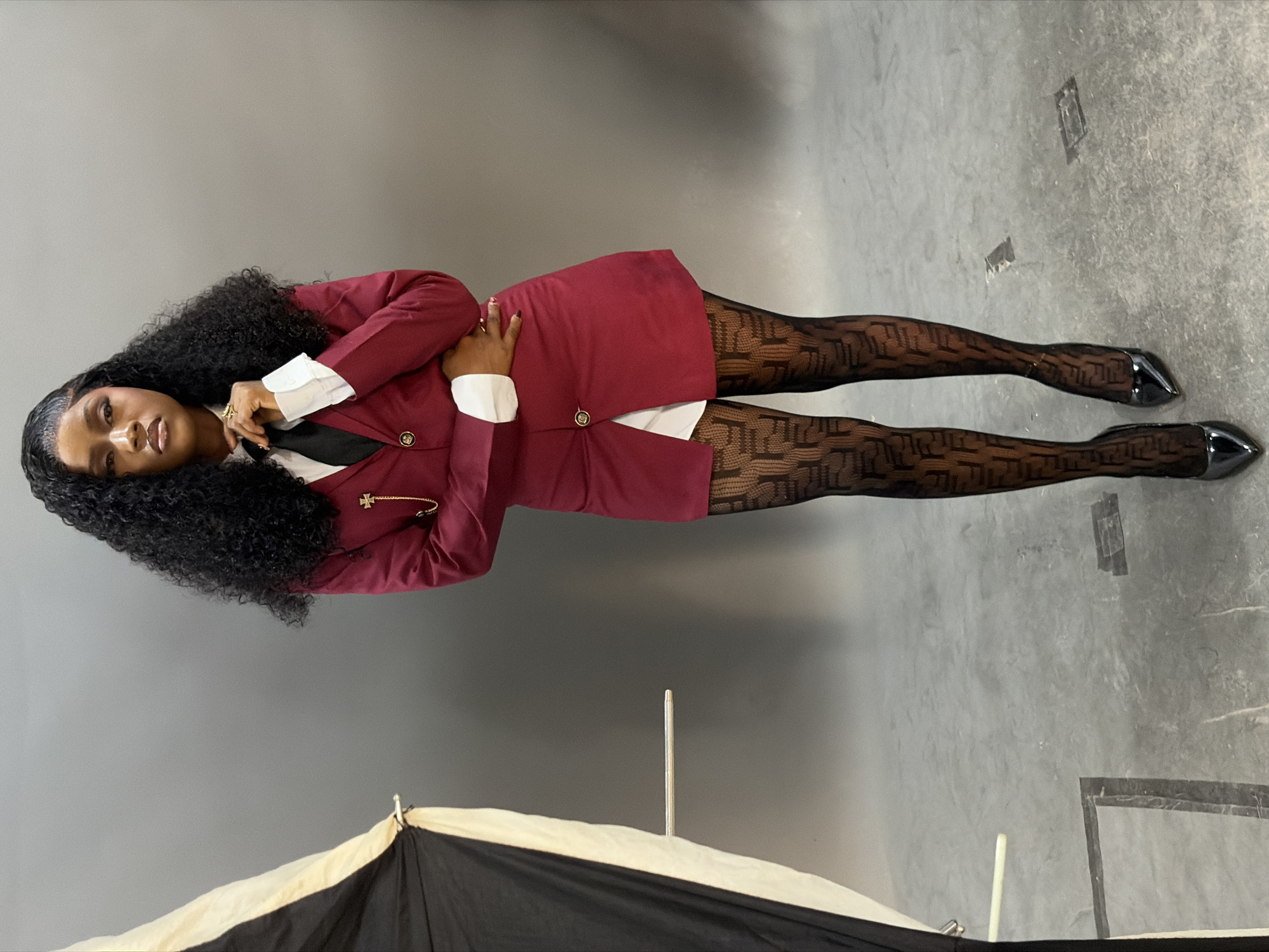 An indoor, full-body portrait captures a Black woman standing centrally in what appears to be a professional photo studio. She has long, dark, curly hair with a side part, and is looking directly at the camera with a neutral expression. Her attire consists of a white collared shirt with prominent cuffs, a black tie, and a maroon blazer-style garment worn as a dress, featuring gold buttons and a gold chain brooch with a cross-like emblem on the left lapel. She is wearing sheer black tights with a repeating monogram pattern and black patent leather pointed-toe high heels. Gold rings adorn her fingers, and dark nail polish is visible. Her left hand is raised towards her collar, while her right hand rests on her hip.
The background is a uniform grey seamless backdrop, transitioning to a light grey concrete floor with some subtle discoloration and markings, including what appear to be taped sections. On the far left, a portion of a black and off-white photographic umbrella diffuser with an extending metallic rod is visible, indicating a studio setup. No discernible features within the image allow for the identification of a specific city, such as Gura Top, Nigeria, or any other geographical location.
