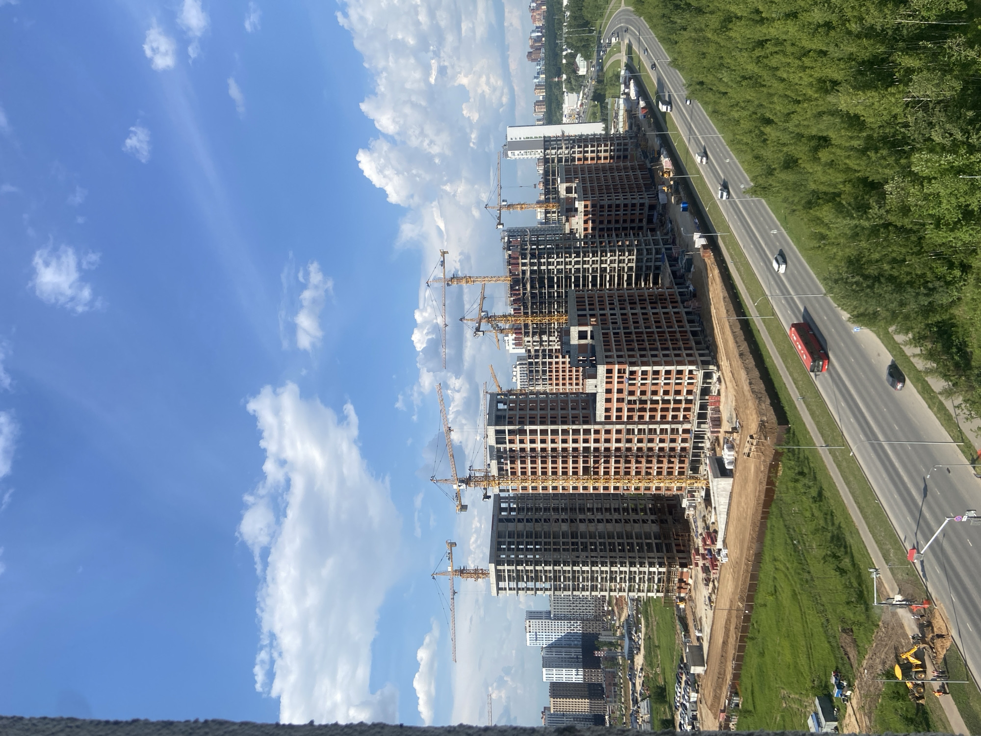 The image depicts a sprawling construction site in Kazan, Russia, with multiple high-rise buildings under various stages of development. A prominent feature is the presence of numerous yellow construction cranes towering over the unfinished structures. The buildings themselves appear to be primarily residential, constructed with concrete frames and brickwork, many of which are still in their skeletal form.
In the foreground, a busy highway curves through the scene, with several vehicles including cars, trucks, and a red bus in motion. Adjacent to the highway is a dense forest with lush green trees, suggesting the construction site is located on the outskirts of the city or in a developing suburban area. Near the construction site, on the left side of the image, there is a patch of green grass with some construction equipment, including a yellow excavator, and several figures, likely construction workers, can be observed. Further in the background, more completed residential buildings and a distant cityscape are visible, indicating the presence of an established urban area. The sky is a clear blue, dotted with scattered white clouds, suggesting a pleasant day. The overall scene conveys a sense of ongoing development and urban expansion within Kazan.