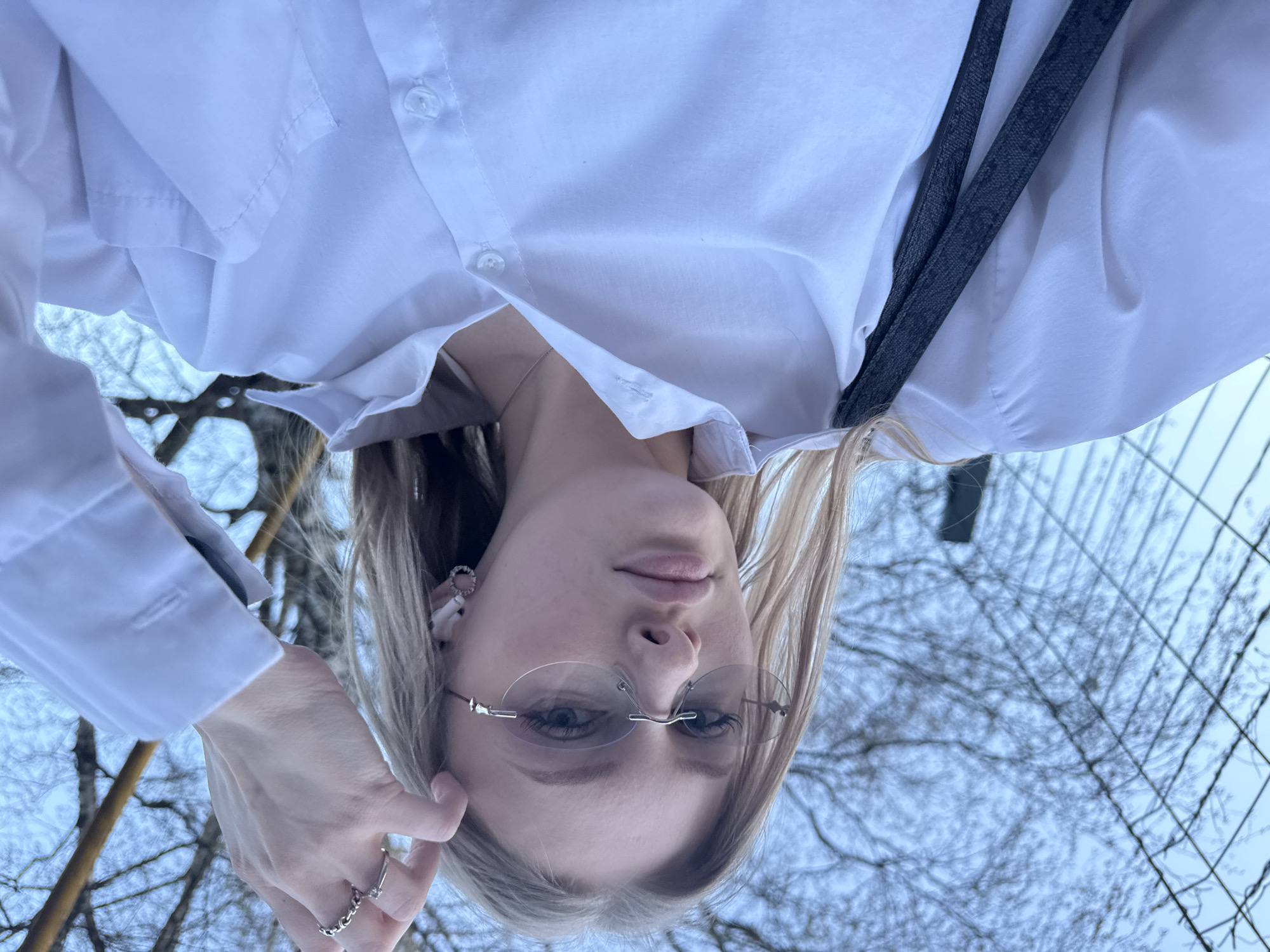 The image shows a blonde woman wearing rimless glasses and a white button-down shirt, looking down at the camera. She is wearing rings on her left hand. A gray strap hangs across her chest. The background shows bare tree branches against a light blue sky. Considering the context clue and the image's features, it's likely that the photo was taken in Perm, Russia, as per the request.