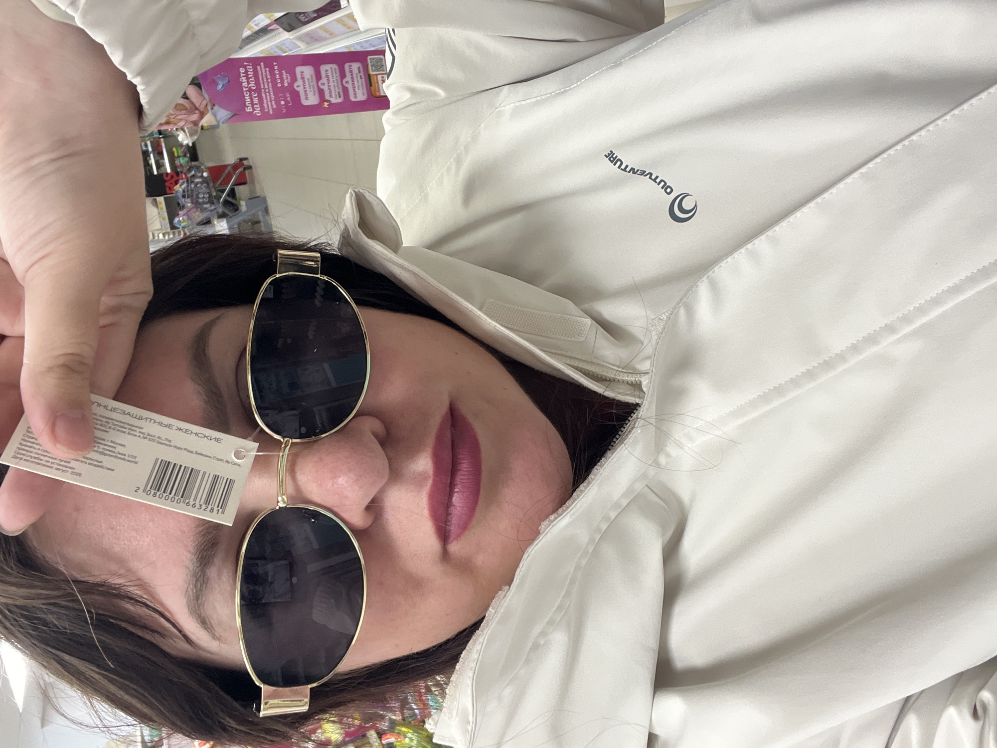 Here is a detailed description of the image:
**People:** The image primarily features a woman. Her face is visible, partially obscured by a pair of gold-rimmed sunglasses with dark lenses and a price tag. She has dark hair and is wearing makeup, specifically lipstick.
**Objects:** The main objects include the sunglasses, a price tag with the text "СОЛНЦЕЗАЩИТНЫЕ ЖЕНСКИЕ" (SUN PROTECTIVE FEMALE), and a light-colored jacket or coat with the brand "OUTVENTURE" visible on the chest.
**Environment:** The image appears to be taken indoors, possibly in a retail setting. There are hints of a store shelf or display in the background.
**Location:** The price tag on the sunglasses shows text in Russian, including the city of Moscow and Yi Wu City, China, which is not Noviy, Russia.