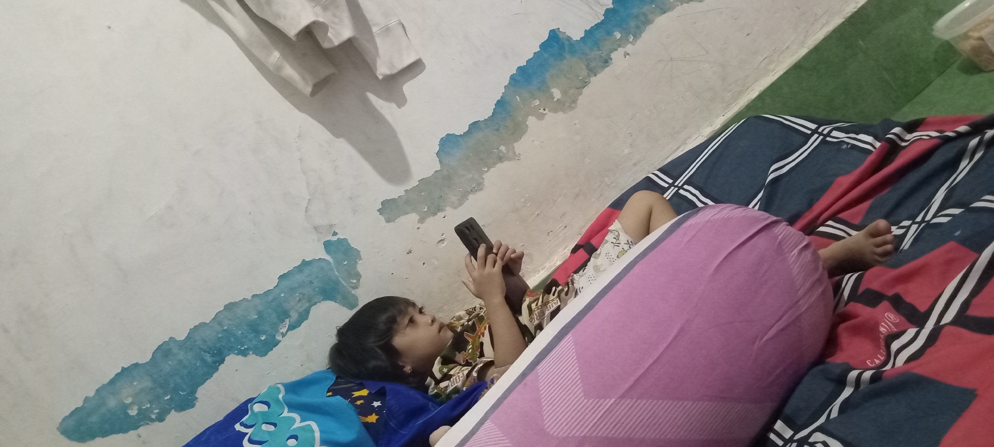 An undated, outdoor, eye-level, medium shot shows a young child lying on their side on a patterned bedspread, looking at a mobile phone held in their hands. The child's head is tilted upwards as they look at the screen of the phone, which is dark. The child is wearing a patterned shirt and has dark hair. Behind the child, a large pink pillow is visible, and the bedspread has a geometric pattern of dark blue, red, and white lines. To the left of the bed, a white wall has peeling blue paint. In the background, a green carpet is visible. The scene appears to be in an indoor setting, possibly a bedroom. There are no identifying landmarks or contextual clues to suggest a specific location within the city of Unknown, Unknown.