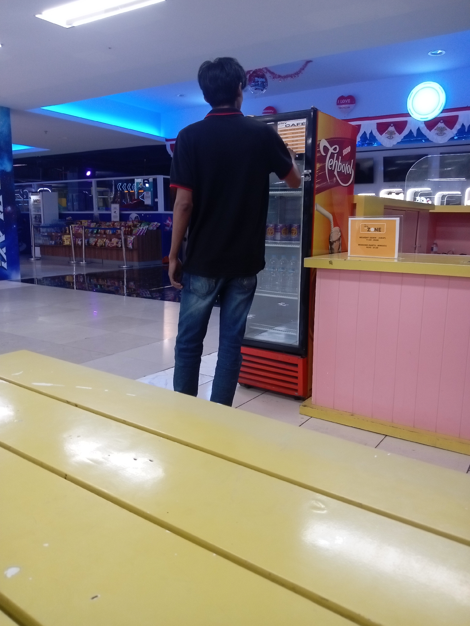 An adult male, viewed from behind, stands facing a commercial refrigerator and a pink counter in what appears to be a food court or a recreational area. He is wearing a black t-shirt with a red collar and blue jeans. The refrigerator, branded "Teh Botol," displays a menu on its front. To the right of the refrigerator is a pink counter with a yellow trim, behind which is a sign that reads "ZONE." In the background, a shelf stocked with various snacks is visible, and above, blue LED lighting illuminates the ceiling. A yellow table is prominently featured in the foreground. The scene is indoors, likely in a public space in Batam, Indonesia, judging by the "Teh Botol" brand which is popular there.