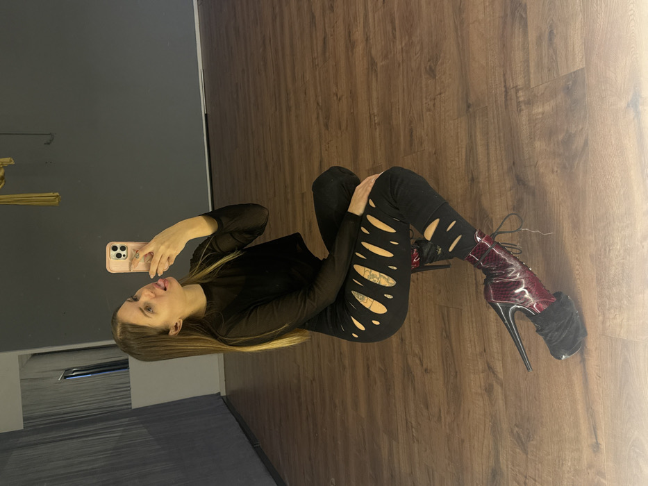 The scene depicts an indoor mirror selfie taken by a female subject within a studio-like environment in Batumi, Georgia. The subject, a light-skinned female with long, straight light brown hair, is crouched or squatting. She holds a light pink smartphone with a visible camera array in her right hand, pointing it towards her reflection in a mirror (not directly visible). Her left arm rests on her left knee, and her expression includes her tongue slightly extended. She is dressed in a black, sheer, long-sleeved crop top and form-fitting black pants with multiple horizontal and diagonal cutouts exposing skin and a visible multi-colored tattoo on her left thigh. She wears dark red/burgundy, high-heeled platform ankle boots with black laces, a prominent black platform, and a slender black heel. A black fur or velvet-like cuff is present at the top of the boots. The room features polished medium-brown wood laminate or hardwood flooring and solid dark grey walls. In the background, a reflection of a dark area is visible within a mirror to the left, and a folded golden fabric element hangs from the ceiling to the right.
