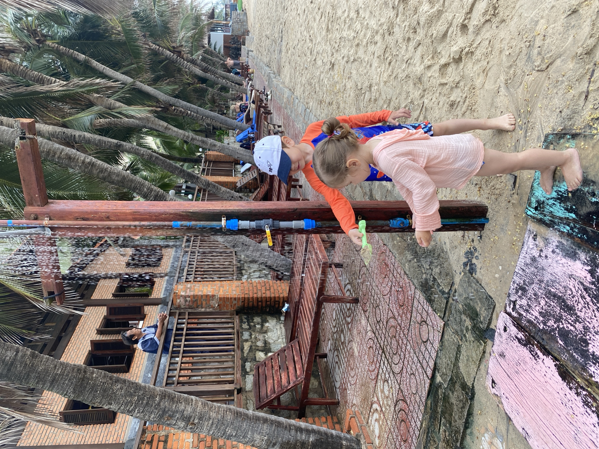 Two children stand next to each other on a beach near Phu Thuy Ward, Vietnam. The child on the left, wearing a blue and orange long-sleeved shirt and a white and blue baseball cap, peers down. The child on the right, wearing a light pink long-sleeved shirt, also looks down. Both children are near a wooden railing with a PVC pipe running along its length. Water is spraying from the pipe. Behind the children is a sandy beach and rows of palm trees. A building with brick and wood construction is visible on the left. The ground is covered with patterned tiles and wooden planks.