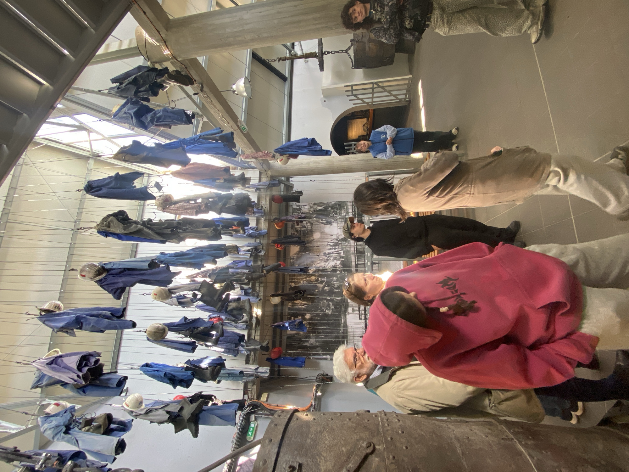 The image shows an interior view of a building in La Motte-d'Aveillans, France. An art installation hangs from the ceiling featuring blue work clothes, hats, and boots suspended on wires. Several people stand on the floor below, observing the installation. Some are wearing casual clothing like hoodies, while others wear jackets. A large, rusty metal object sits in the foreground. A historical photograph or display is visible in the background. The building appears to be a museum or exhibit space.