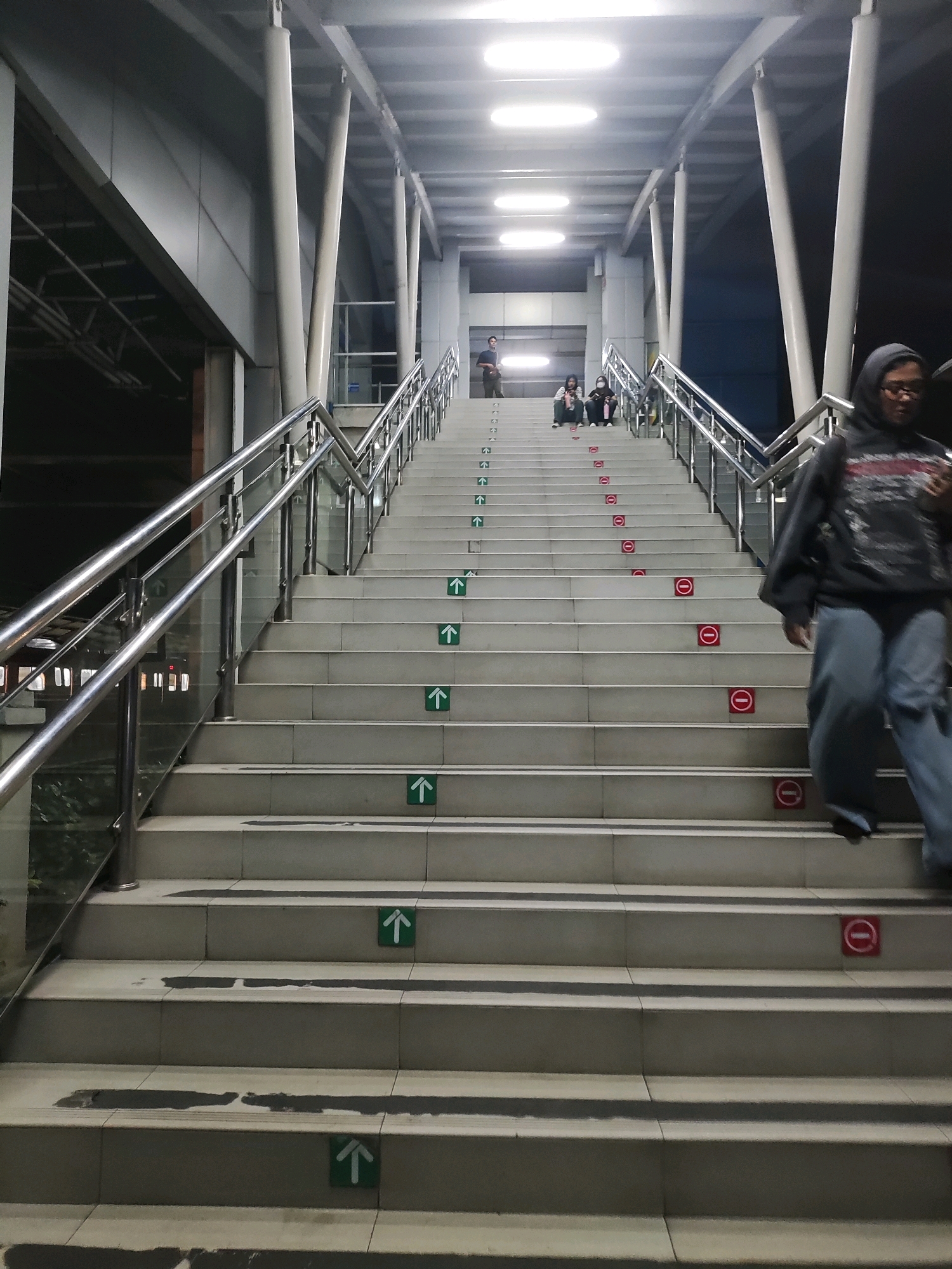 The image depicts a wide, outdoor staircase with a metal railing on either side. The staircase is made of light-colored concrete tiles. Several green arrows pointing upwards are placed on the steps, indicating the direction of ascent. Red circular signs with a horizontal line through them, signifying "do not enter" or "prohibited," are also present on some steps, alternating with the arrows.
At the top of the staircase, several people are visible sitting or standing. Further down, a person wearing a dark hoodie and blue pants is walking down the stairs, with their face blurred. The staircase is part of an elevated walkway or bridge, with a covered ceiling above supported by metal columns. The overall environment appears to be a public transportation hub or a pedestrian overpass in an urban setting.
The image is located in East Jakarta, Indonesia. Specifically, this staircase is located in the vicinity of the Cikini Station, a railway station in Central Jakarta, Indonesia, which is part of the greater Jakarta metropolitan area.