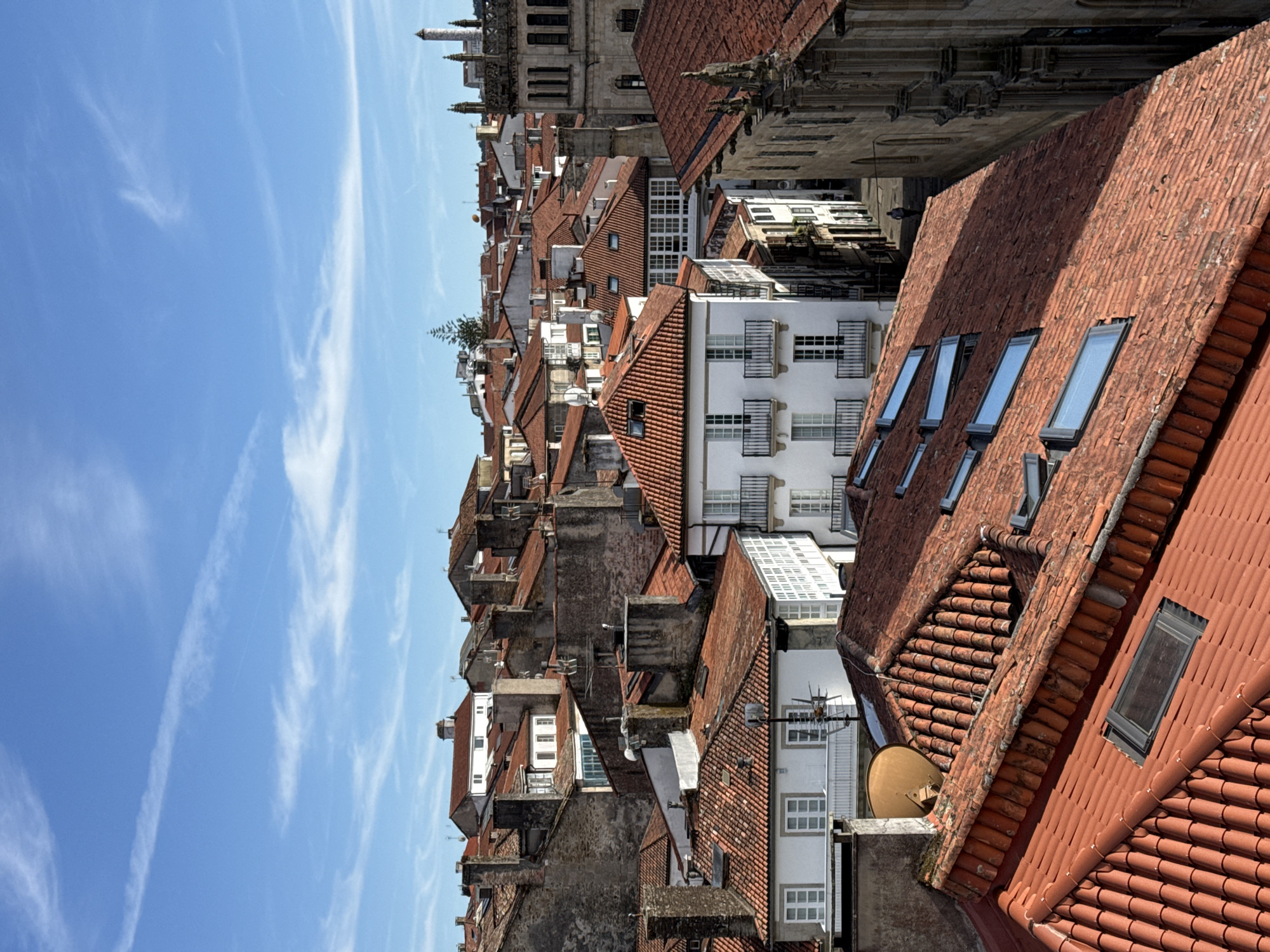 The image depicts a view of the rooftops of the old town of Santiago de Compostela, Spain, under a bright blue sky with scattered white clouds. The foreground is dominated by a close-up of weathered, terracotta tiled roofs, some with skylights. In the mid-ground, more buildings with red tiled roofs and white facades are visible, interspersed with older stone structures and chimneys. The architecture suggests a historic European city. The background reveals a denser collection of buildings and a glimpse of a tall, ornate stone tower, possibly a cathedral or church spire, characteristic of the city's historical center. The lighting suggests it is daytime, likely late morning or early afternoon, casting distinct shadows on the rooftops and buildings. A narrow street is partially visible on the right, with a lone figure walking away from the camera. The overall scene conveys a sense of timeless urban landscape.