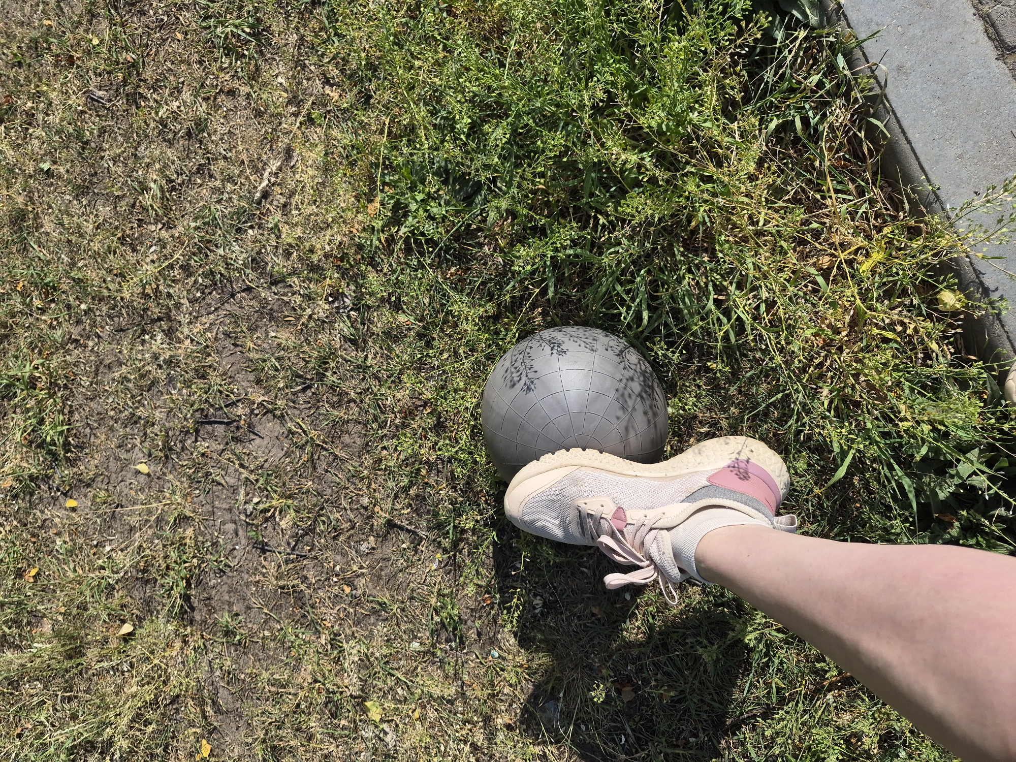A close-up shot features a person's leg and foot wearing a pink and white sneaker, resting on a gray exercise ball. The ball and foot are situated on a patch of mixed grass and dirt, next to a concrete curb. The scene appears to be outdoors, possibly in a park or green space. The location is specified as Unknown, Unknown.