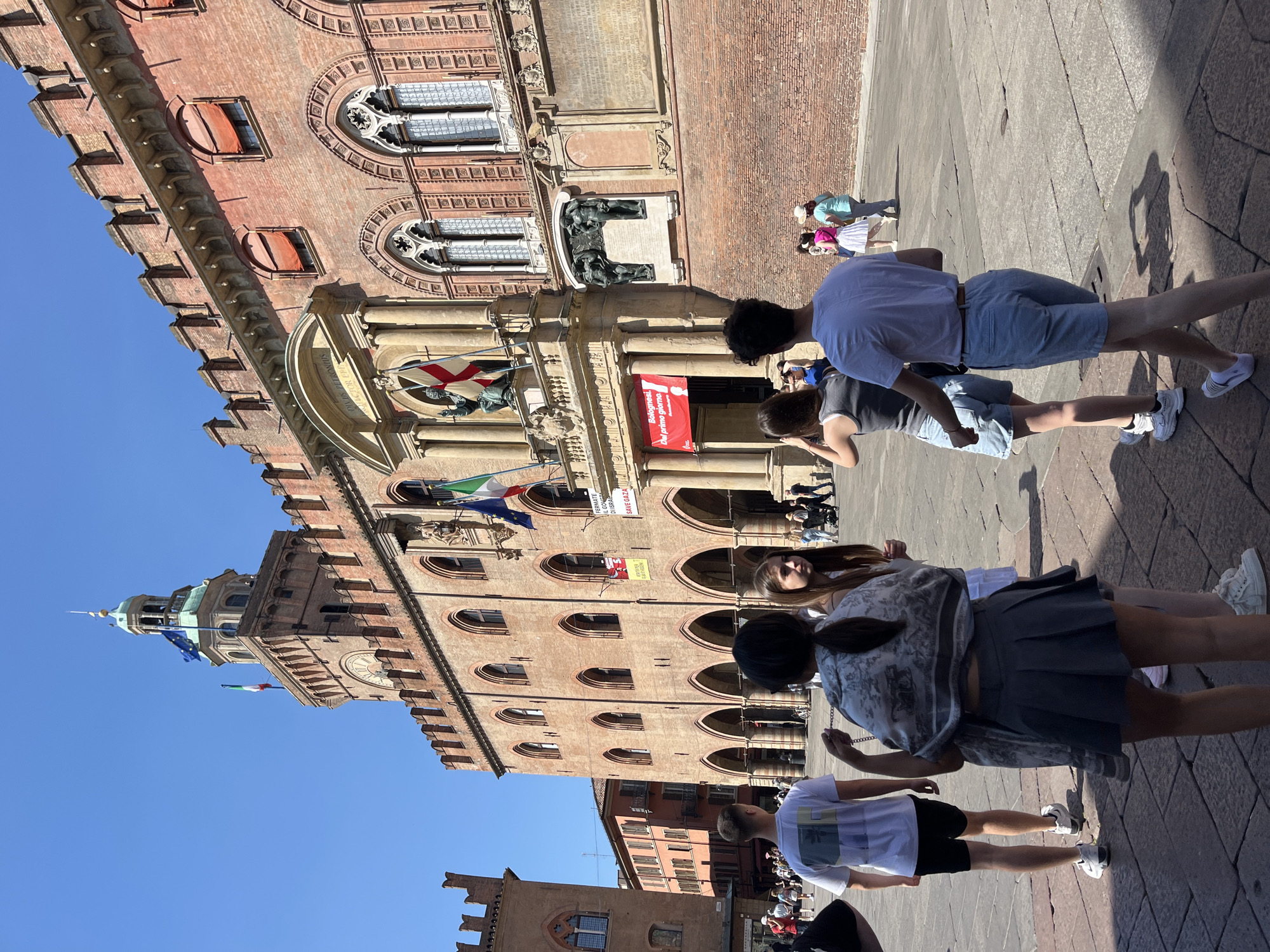 The image shows a daytime view of Piazza Maggiore in Bologna, Italy, with the Palazzo d'Accursio (also known as Palazzo Comunale) as the focal point. Several people are visible, primarily in the foreground, some seemingly taking photos of the building. The Palazzo d'Accursio is a large, historical brick building with multiple levels, arched windows, and flags displayed. The sky is clear and blue. The paving stones of the piazza are visible on the ground.