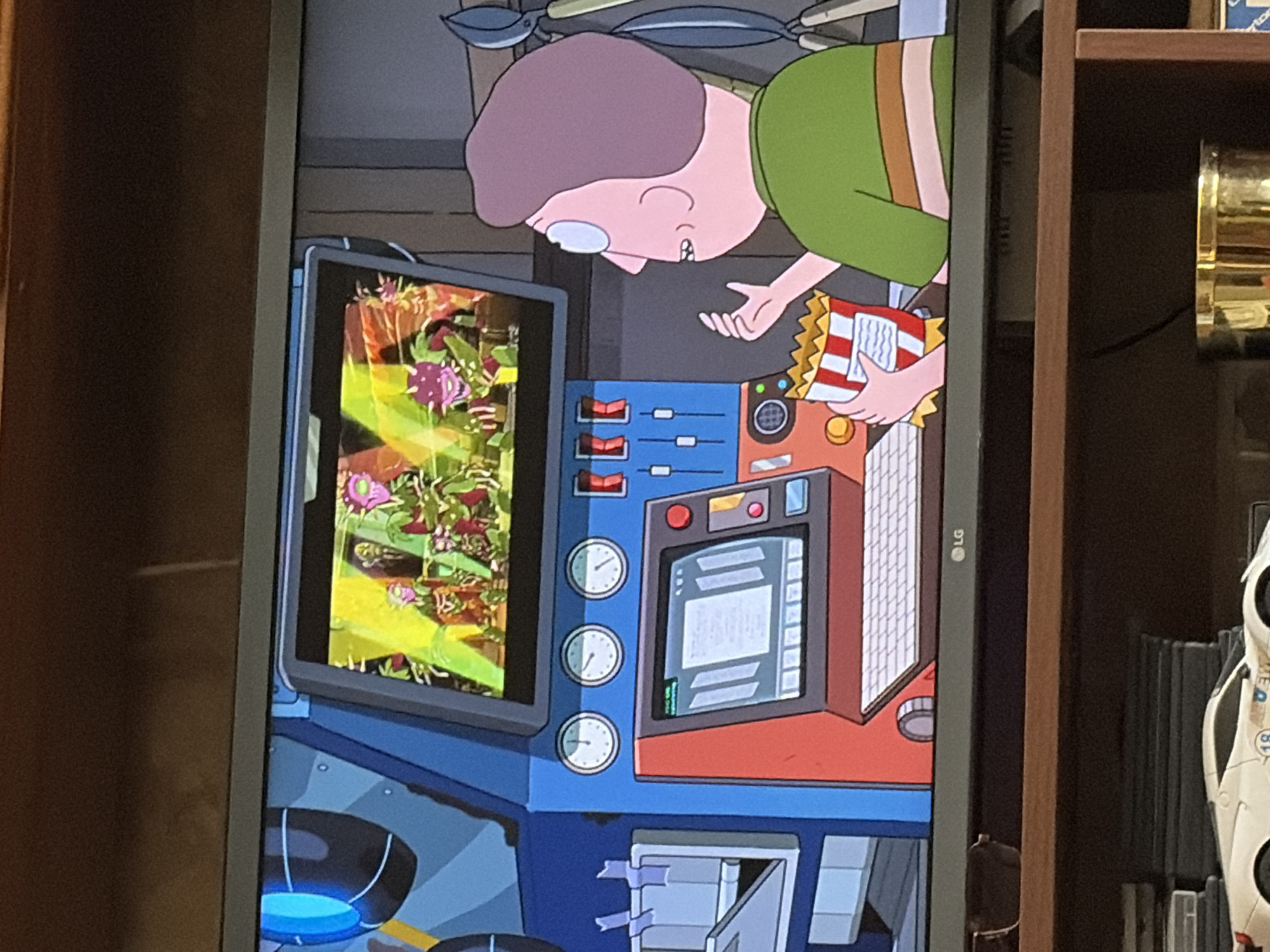The image shows a television screen displaying a cartoon scene from Rick and Morty. On the screen, several small, purple, dinosaur-like creatures are visible in a bright, colorful environment. In the foreground, to the right of the television, a character resembling Jerry Smith from the show is seen from the side. He is wearing a green shirt with brown and pink stripes and is holding a box of popcorn. The television is placed on a wooden shelf, with other items visible on the shelf below, including a white toy car and a golden container. The background of the television screen shows a complex blue and red machine with various buttons, dials, and a keyboard.