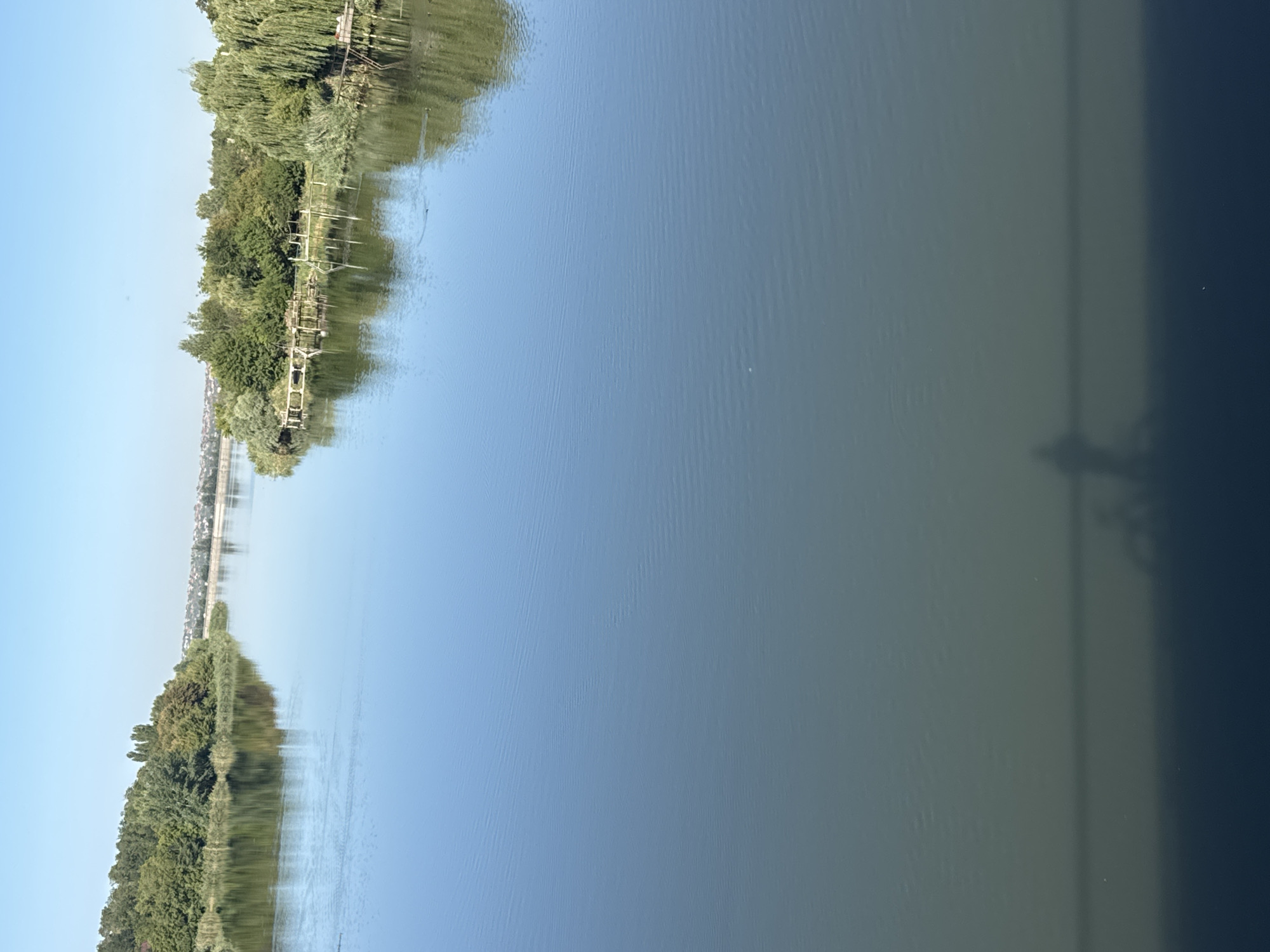 This image shows a wide river in Rostov-on-Don, Russia, on a clear day. The water is calm, reflecting the blue sky and the lush green trees lining the riverbanks. On the left bank, dense foliage creates a verdant wall, while the right bank features a more varied landscape with trees, reeds, and wooden structures resembling piers or fishing platforms. In the distance, a hazy cityscape with buildings is visible on the horizon. The foreground of the image shows a dark shadow cast on the water, which appears to be the silhouette of a person on a bicycle, possibly viewed from a bridge or elevated vantage point. The overall scene conveys a sense of tranquility and natural beauty within an urban setting.
