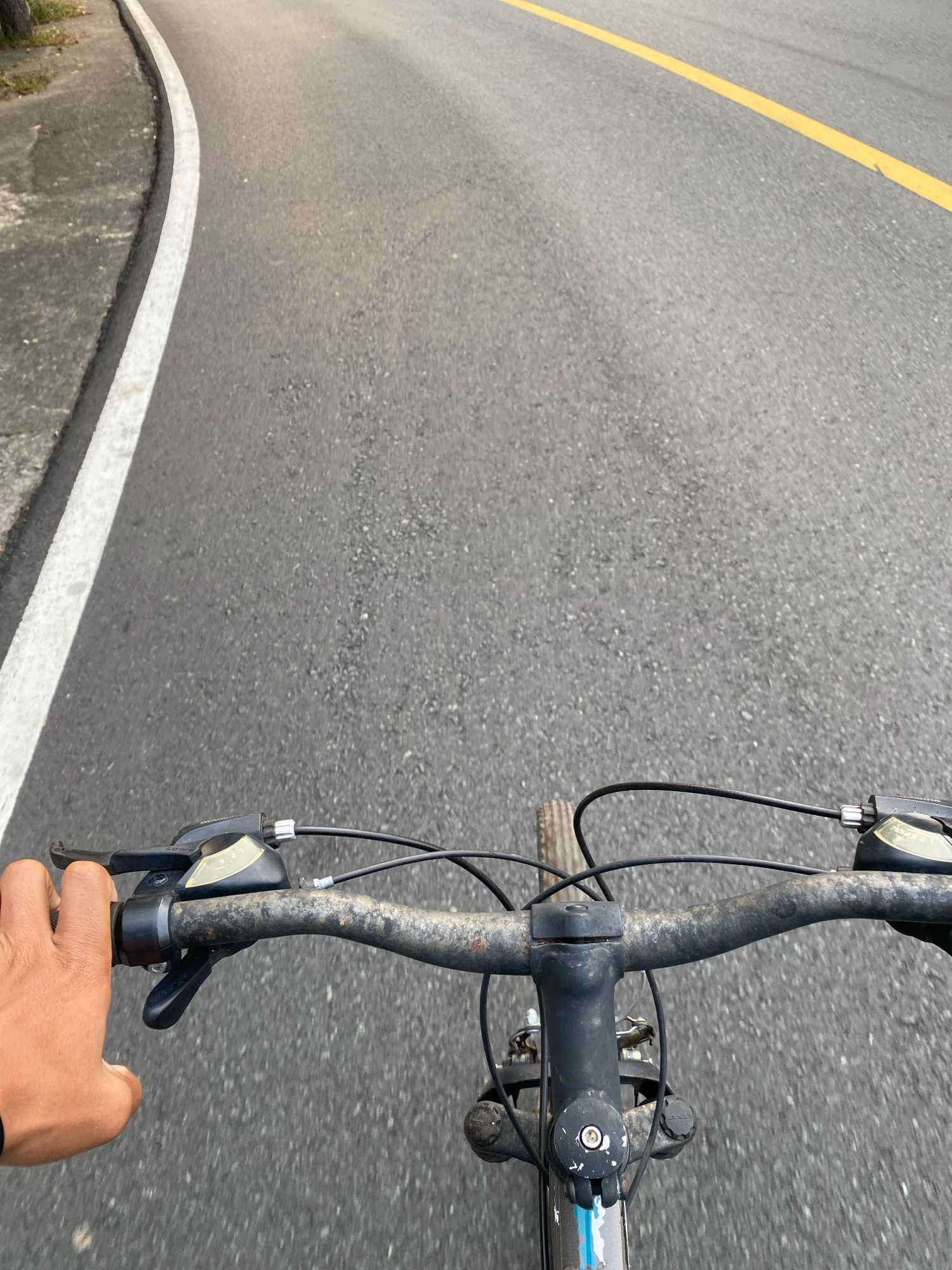 An upward-angle, first-person perspective shot shows a hand gripping the handlebar of a bicycle as it rides along a paved road. The road is marked with a solid white line on the left and a broken yellow line on the right, suggesting it is a two-lane road. The asphalt surface shows a rough texture with scattered small stones. The bicycle's handlebars are dark gray and show signs of wear and rust. A gear shifter is visible on the right handlebar. The background is mostly the road, with a hint of vegetation on the far left, bordering the road. The lighting suggests it is either early morning or late afternoon. The scene is likely set in Tanjung Pinang, Indonesia, given the characteristic road markings and typical urban landscape of the region.