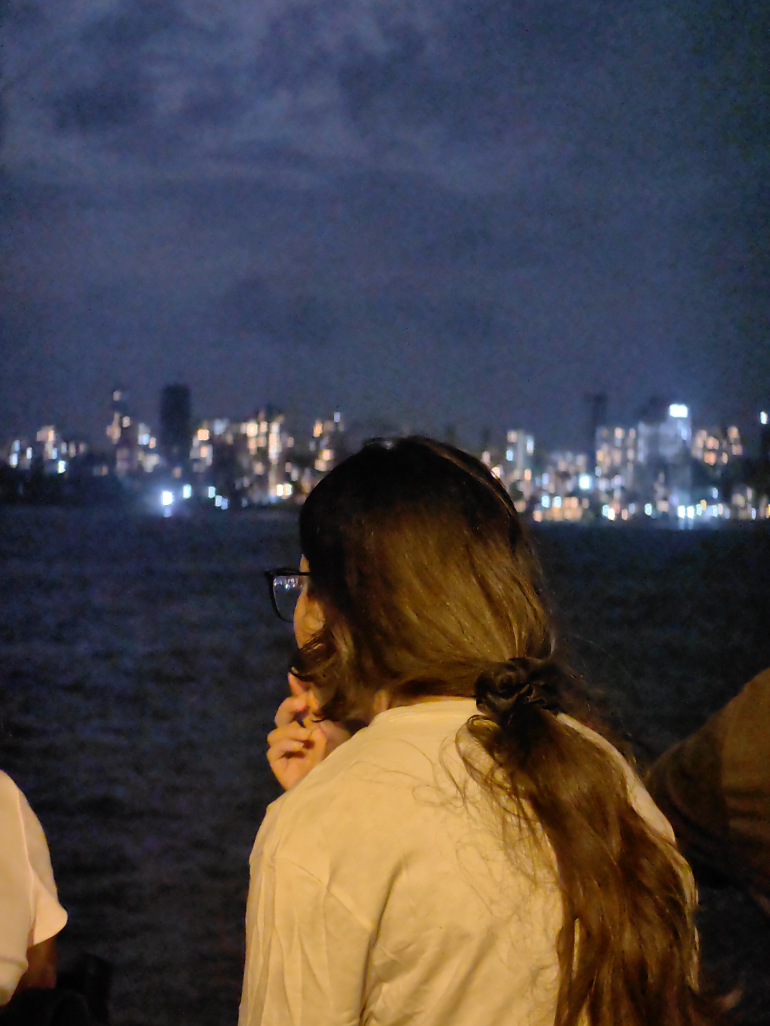 A woman with long, dark hair tied in a ponytail is seen from behind, looking out at a city skyline at night, which is likely Mumbai, India, given the context. She is wearing glasses and a light-colored shirt. The city lights are blurred in the background, creating a bokeh effect over the water. The sky is dark blue, suggesting it is nighttime. Another person, partially visible, is to the left, wearing a white shirt. A third person's shoulder and back are visible on the right, wearing a brown garment. The overall scene is dark, with the city lights being the most prominent source of illumination.