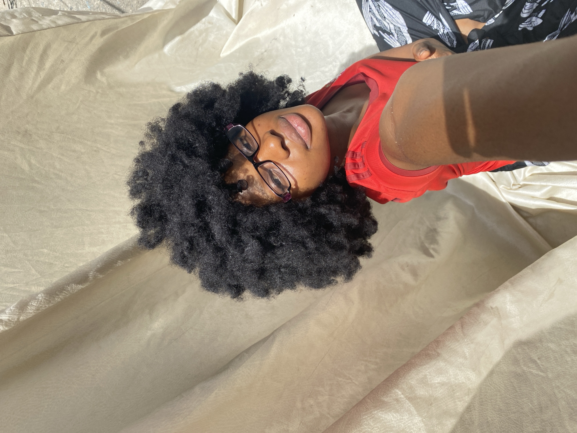 A person lies on a light tan surface, presumably fabric, in an outdoor location with sunlight. The person is wearing a red shirt and black and white patterned pants. The person's dark hair is large and textured, and they are wearing glasses. Their eyes are closed. The location is Iranla, Nigeria.