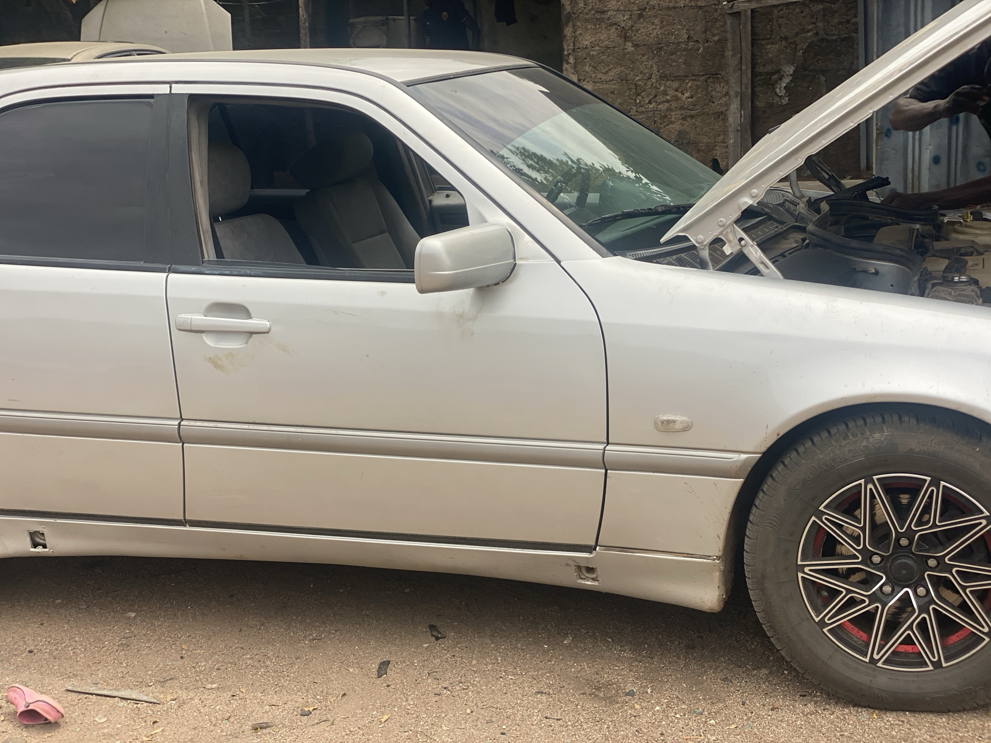 An exterior, eye-level, medium shot shows the driver's side of a silver Mercedes-Benz sedan parked on a dirt lot in Jalingo, Nigeria. The car's hood is open, revealing the engine compartment. A Black male, with dark skin and short black hair, is partially visible on the right side of the frame, reaching into the engine bay. The car has tinted windows, silver side mirrors, and a custom black and red rim on the front driver's side wheel. The ground is covered in loose dirt and gravel, with a pink flip-flop visible in the lower left corner. The background consists of a textured, light brown brick wall and some greenery. The scene appears to be an informal mechanic's bay.