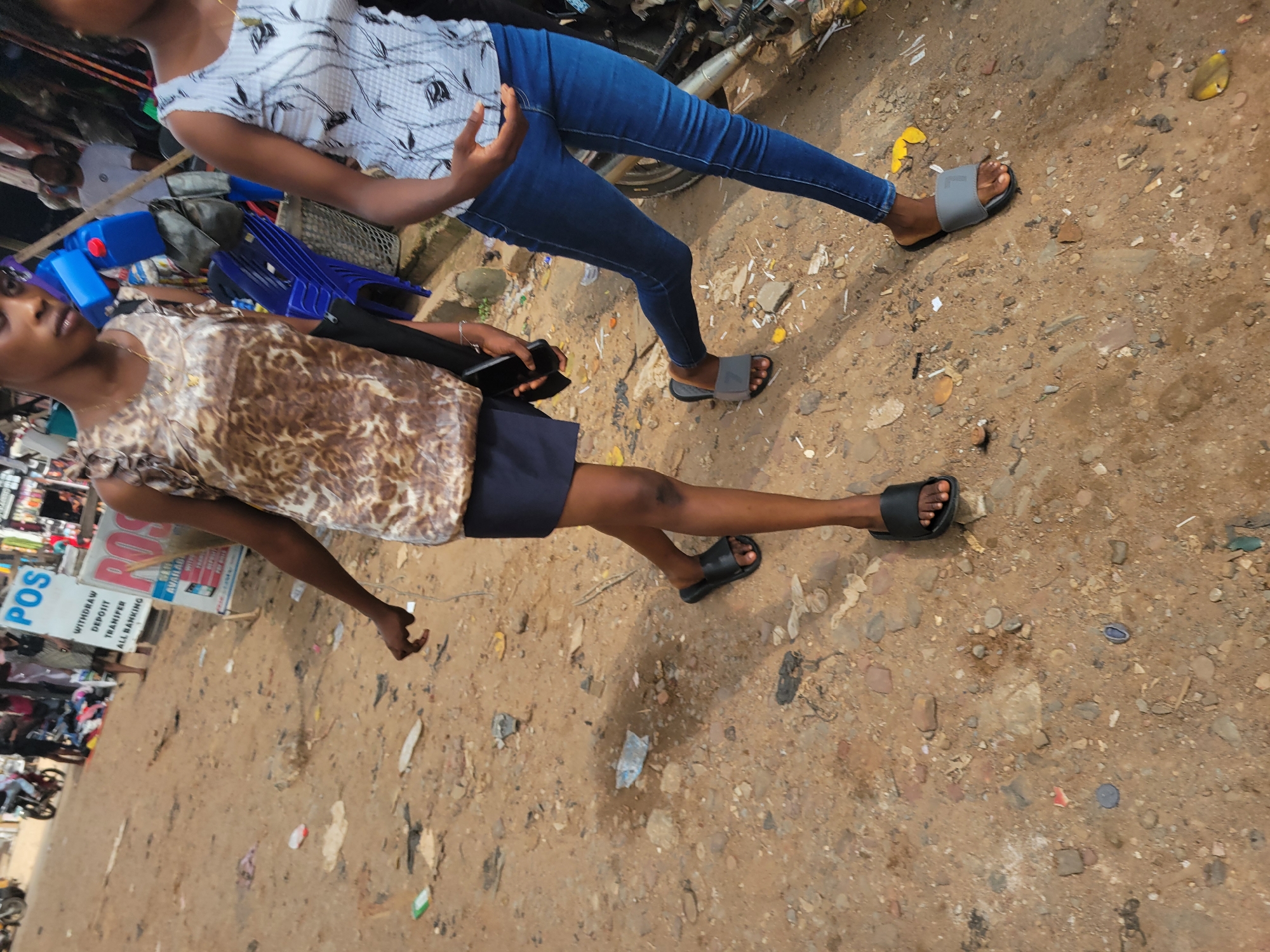 Two young women are standing on a dirt path, likely in Egbagu, Nigeria, based on the location context. The woman in the foreground is wearing a brown and white leopard print top and a dark blue skirt. She is holding a black smartphone in her right hand and wearing black slide sandals. She has dark skin and dark hair, and appears to be looking downwards.
The woman behind her is wearing a white and black patterned t-shirt and blue jeans. She is also wearing grey slide sandals. Her body is angled away from the camera, and her right arm is raised, with her hand visible.
The ground is a mixture of dirt, small stones, and scattered debris. In the background, there are glimpses of various objects including a blue plastic chair and some red and blue containers. To the left of the foreground woman, there is a white sign with red lettering. Visible text on the sign includes "POS", "WITHDRAW", "DEPOSIT", "TRANSFER", and "ALL BANKING". This suggests a service point or business is nearby.
The lighting and shadows indicate it is daytime, possibly late morning or early afternoon, with the sun casting distinct shadows. The overall scene appears to be an outdoor, somewhat informal setting, possibly a market area or a street with local shops. There are no strong indicators of emotion from either individual, they appear to be in a casual stance.