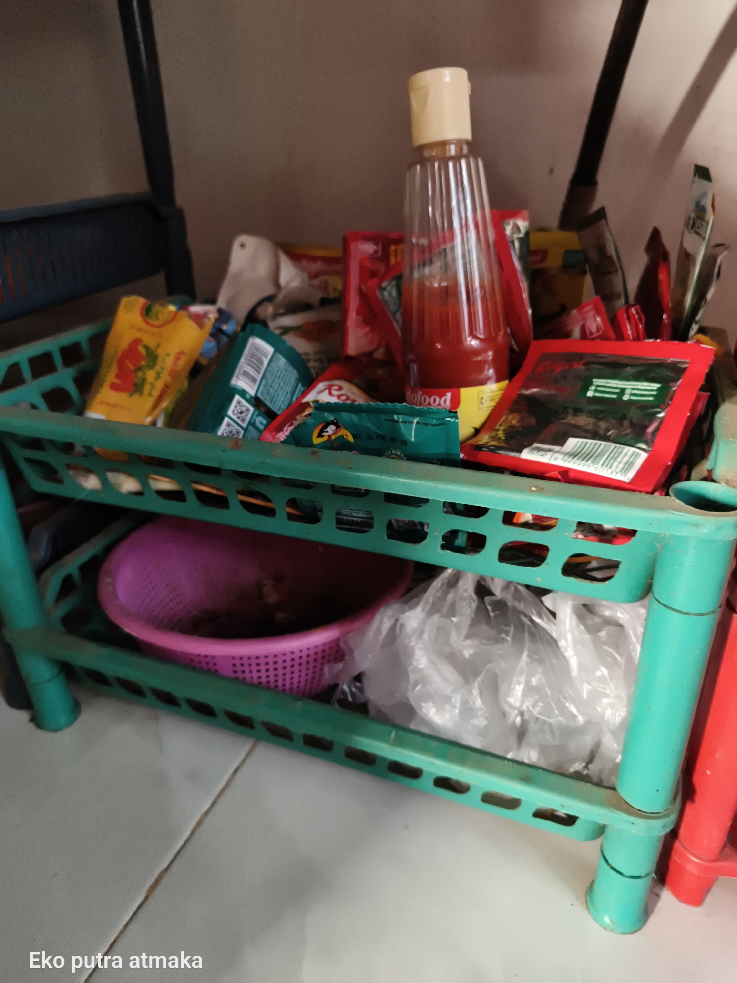 This is a medium shot, indoor, eye-level photograph of a two-tiered green plastic shelving unit filled with various food items and kitchenware. The shelf is situated in what appears to be a pantry or storage area. On the top shelf, there is a tall, clear plastic bottle with a beige cap containing a red sauce, likely chili sauce, positioned in the center. To the left of the sauce bottle are several packets of yellow and red packaging, including what looks like a small bag of seasoning. Behind the sauce bottle, a few other food packets and a white tissue are visible. On the right side of the top shelf, more food packets are arranged.
The bottom shelf of the unit holds a pink plastic colander or sieve, filled with some unidentifiable items, possibly food remnants. To the right of the colander, several clear plastic bags are loosely packed, suggesting storage or leftover food. The shelving unit itself shows signs of wear and some dust. The background is a plain, light-colored wall, and the floor is tiled in a light gray pattern with visible grout lines. The overall impression is one of a functional, if somewhat cluttered, storage space. The bottom left corner of the image contains text that reads "Eko putra atmaka."