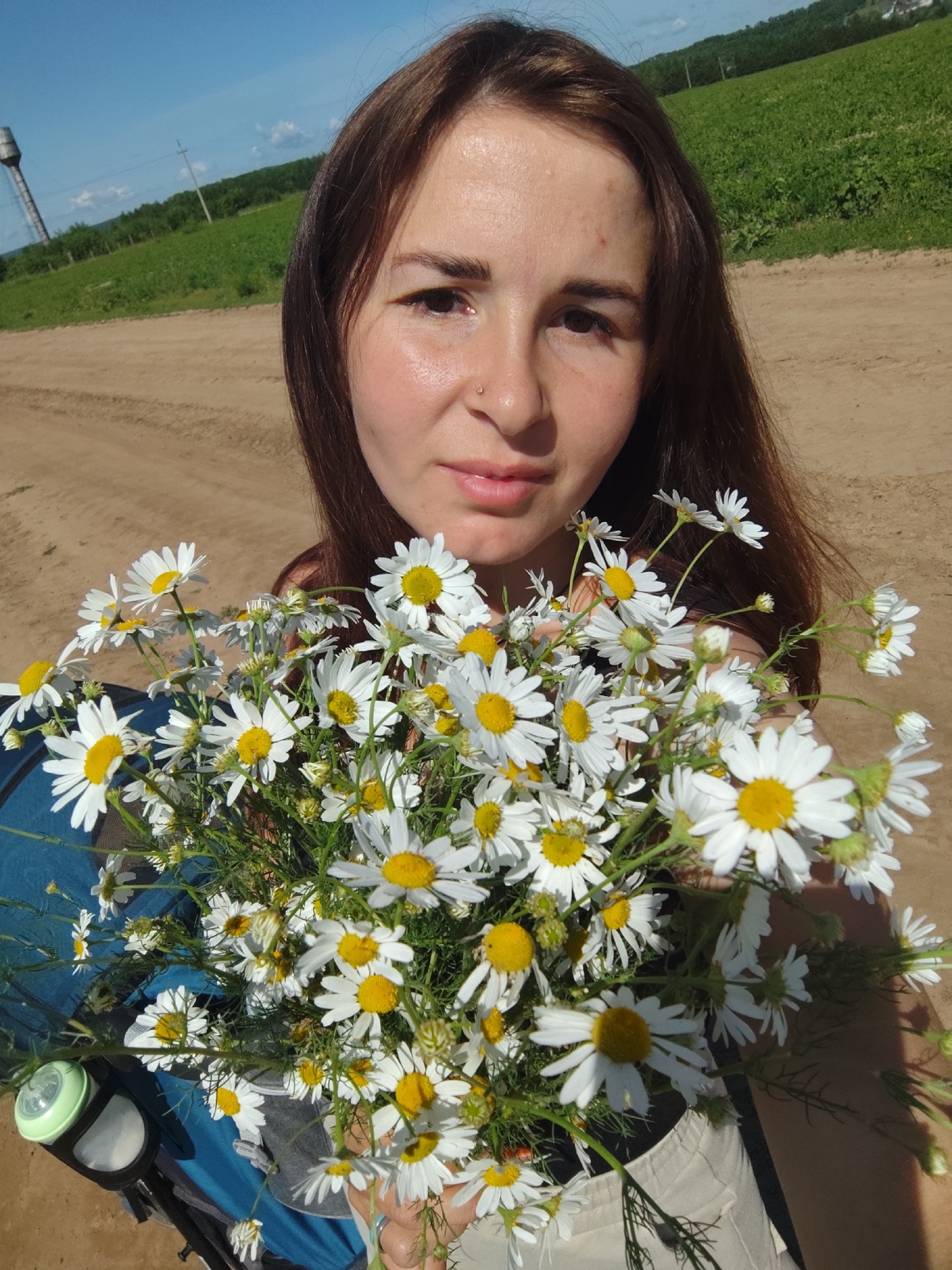 In the city of Foky, Russia, a young woman with brown hair and a fair complexion holds a large bouquet of white daisies with yellow centers. She is positioned in the foreground, looking directly at the camera with a neutral expression. Her face is partially obscured by the flowers. Behind her, a dirt road stretches into the distance, bordered by a green, grassy field. A tall, industrial-looking tower stands on the left side of the field, and trees line the horizon under a clear blue sky with scattered clouds. To the lower left of the woman, a blue object, possibly part of a stroller or carrier, is visible. The overall scene depicts a sunny outdoor setting with a focus on the woman and her floral arrangement.