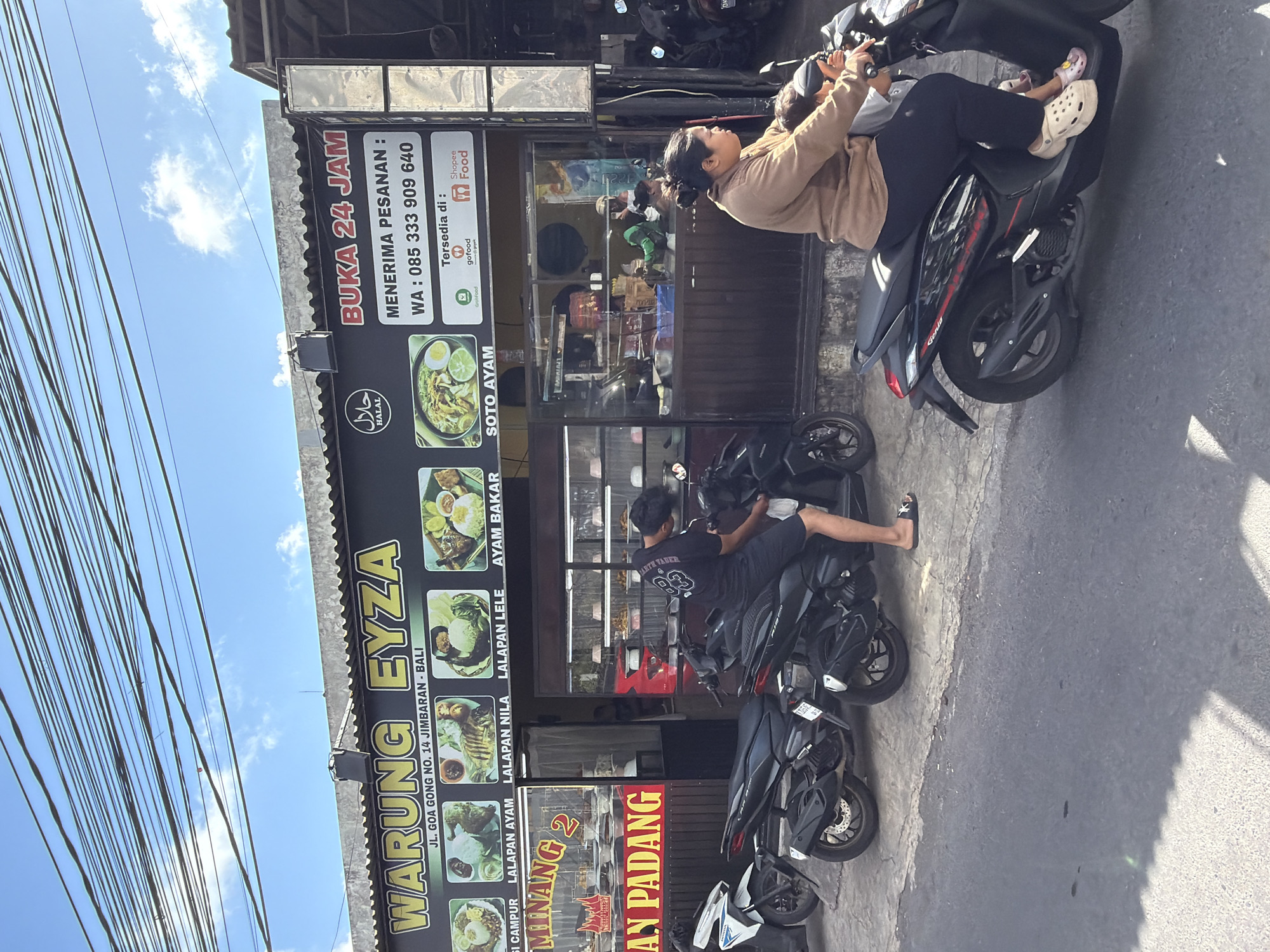 The image depicts the storefront of a restaurant named "WARUNG EYZA" located at JL. GOA GONG NO. 14, JIMBARAN - BALI, Kuta Selatan, Indonesia. The establishment is open 24 hours and accepts orders via WhatsApp at 085 333 909 640. It also lists availability on GoFood and ShopeeFood. Above the entrance, a banner displays images of various Indonesian dishes, including "NASI CAMPUR," "LALAPAN AYAM," "LALAPAN NILA," "LALAPAN LELE," and "AYAM BAKAR." Adjacent to this, there is a section for "MINANG 2" and "AN PADANG," suggesting it also serves Padang cuisine.
In the foreground, several motorcycles are parked along the roadside. A man in a black t-shirt and shorts is interacting with one of the motorcycles, seemingly adjusting something on it. Further in the foreground, a woman is riding a black motorcycle, with a child seated in front of her. She is wearing a brown long-sleeved shirt and dark pants, and the child is wearing a grey shirt. The environment appears to be a street scene with a bright, sunny sky and visible power lines overhead. The building facade is a dark color with yellow lettering for the restaurant name. The interior of the restaurant is visible through the glass windows, showing display shelves with food containers and a few people inside.