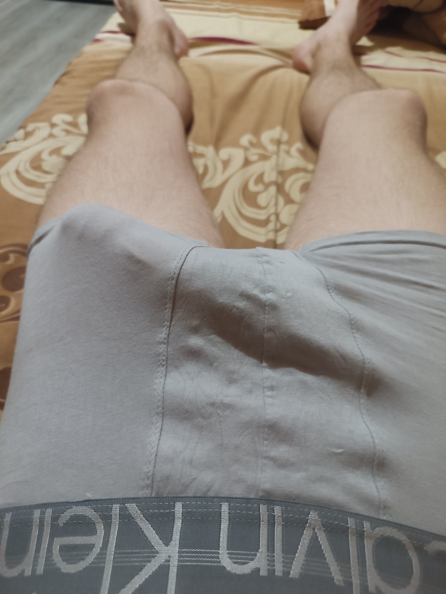 The image shows a close-up, overhead view of a person's legs and groin area, covered by gray Calvin Klein boxer briefs. The person is lying on a bed with a patterned duvet in shades of brown and beige. The lighting suggests it is daytime, possibly indoors. The scene is intimate and casual. Visible text on the boxer briefs reads "CALVIN KLEIN". The focus is on the lower body and the undergarment, with the legs and feet partially visible at the top of the frame. The fabric of the boxer briefs is smooth and appears to be a cotton blend. The background duvet has a swirling, floral-like pattern.