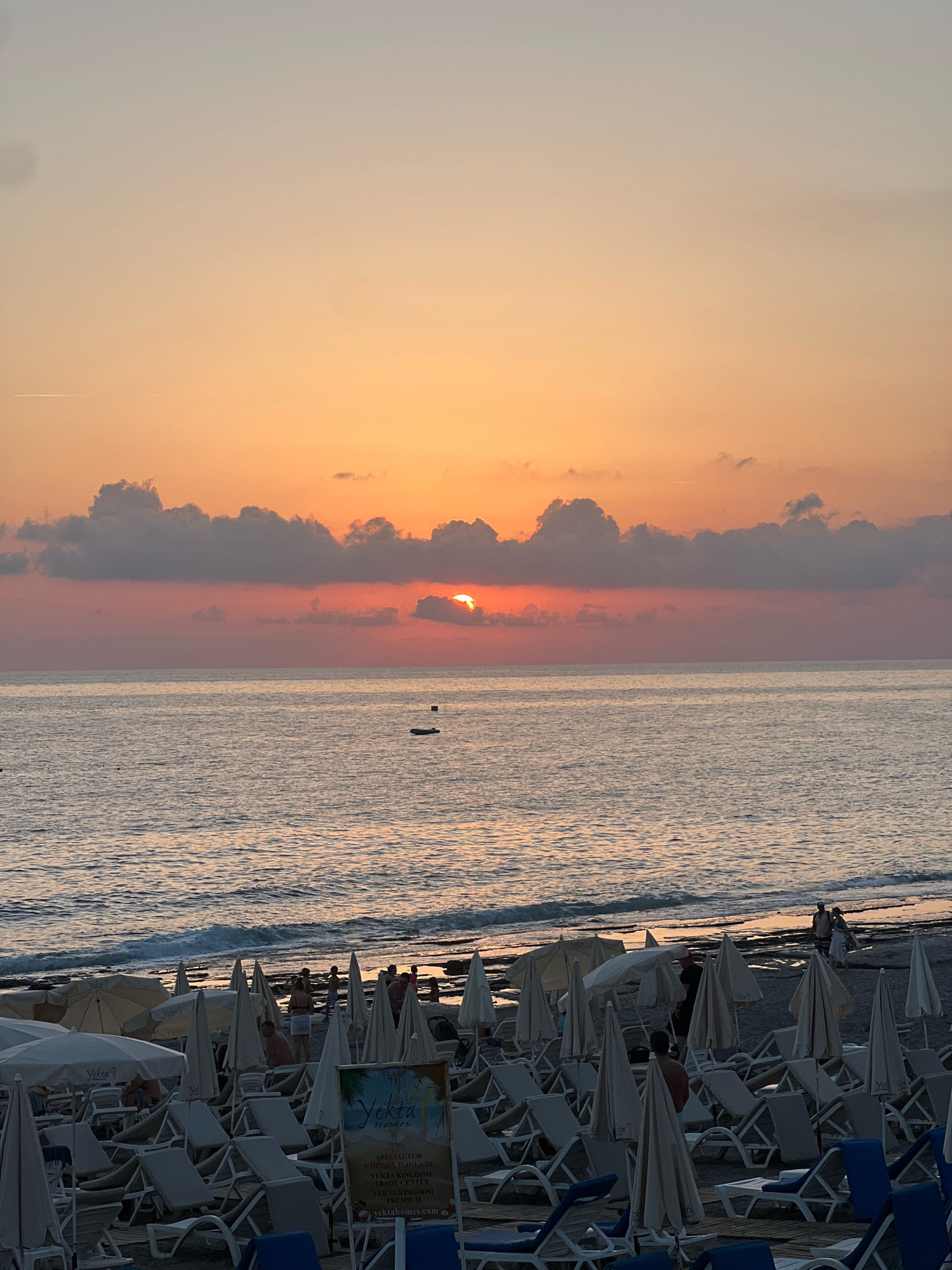 The image depicts a beach scene at sunset in Alanya, Turkey, likely Kleopatra Beach due to the presence of numerous sun loungers and umbrellas. The sun is low on the horizon, partially obscured by clouds, casting a warm orange and pink glow across the sky and the calm sea. Several people are visible on the beach, some walking near the water's edge and others seated on loungers. The foreground is filled with rows of empty sun loungers and closed umbrellas, suggesting the end of the day. A sign with Turkish text and a logo for "Yekta Homes" is visible in the lower right portion of the image. In the distance, a small boat is visible on the water. The overall atmosphere is tranquil and serene.