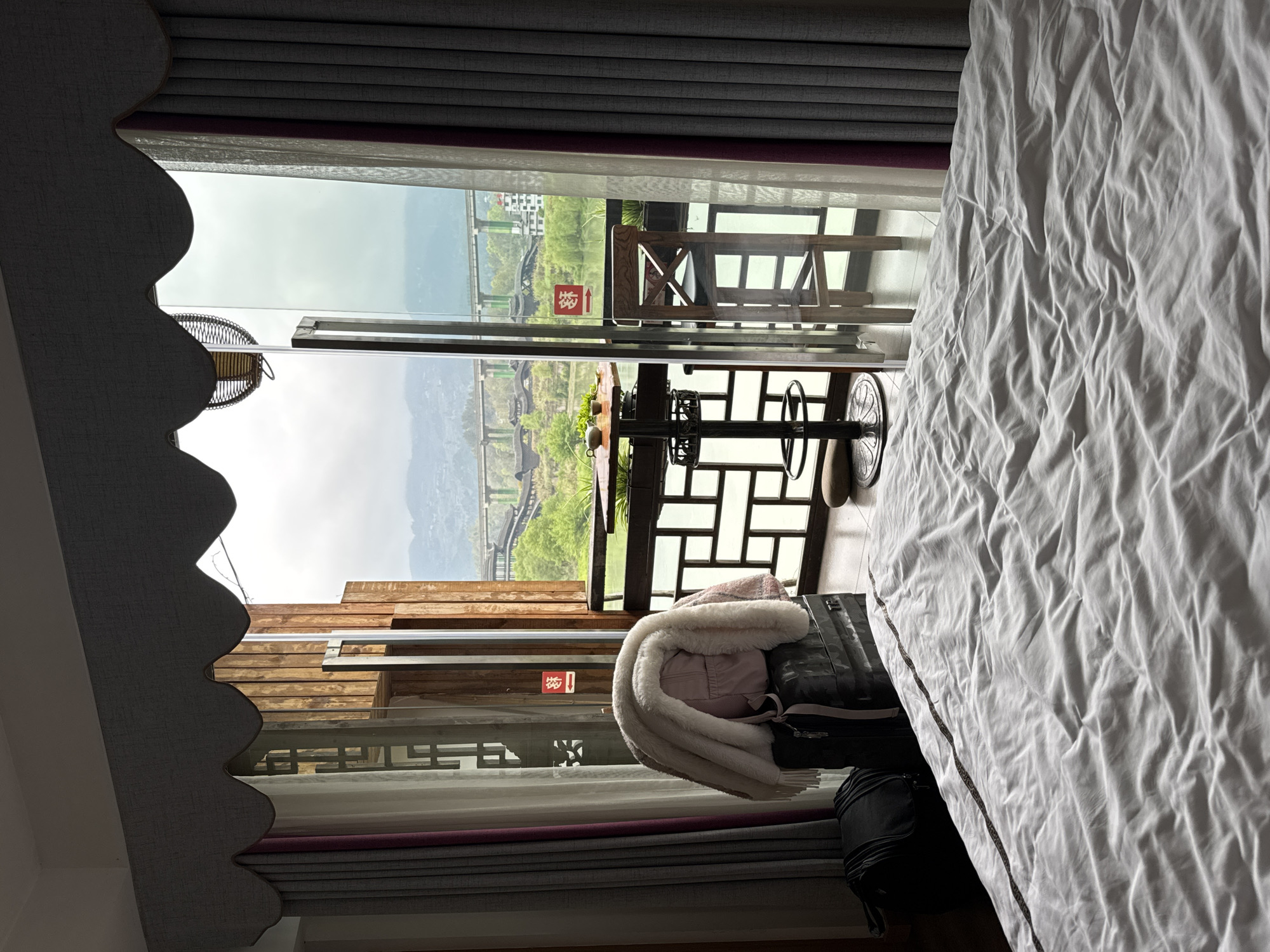 From a bedroom in Zhuoshui, China, a view through a sliding glass door reveals an outdoor table and chairs on a balcony. Beyond the balcony, a hillside is visible with structures like bridges and buildings. Luggage sits stacked in front of the sliding door. The room is furnished with a bed covered in white sheets. The window is framed by grey and purple curtains.