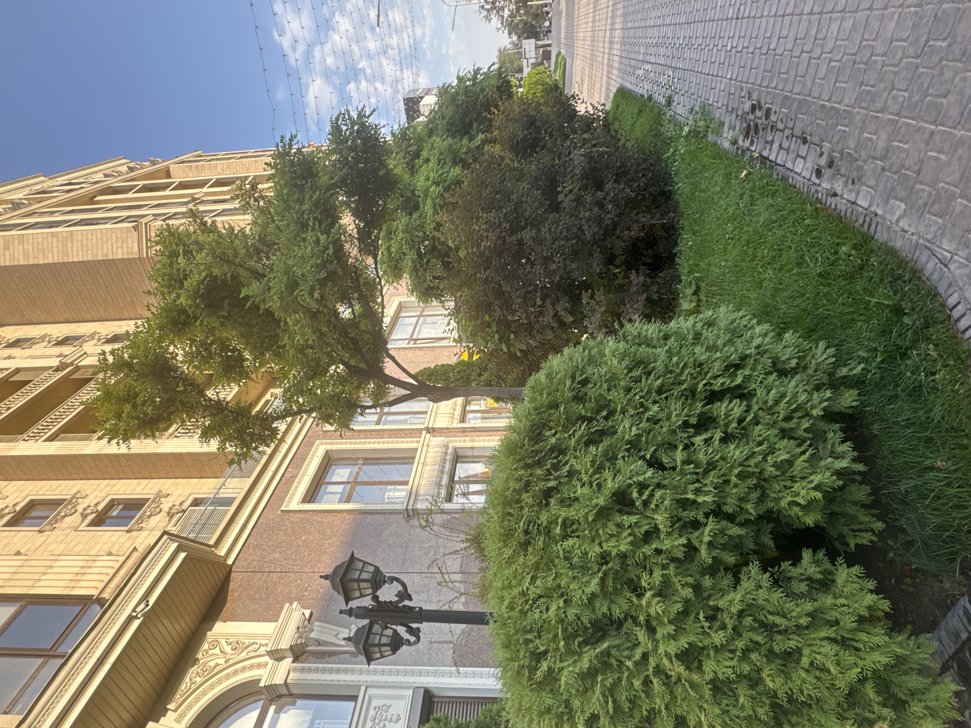 The image shows a paved walkway bordering a manicured garden area in front of a multi-story building. The building appears to be modern, with a light-colored stone facade and arched windows on the lower levels. There are several trees and shrubs in the garden, including a large, dense evergreen bush in the foreground. A lamppost with two lights stands near the evergreen bush. The walkway is made of interlocking paving stones, and a small gutter runs along its edge. In the background, the sky is blue with some clouds, and string lights are visible overhead. The scene suggests an urban environment, possibly a residential or commercial area. Based on the architectural style and landscaping, this location is consistent with areas in Bishkek City, Kyrgyzstan.