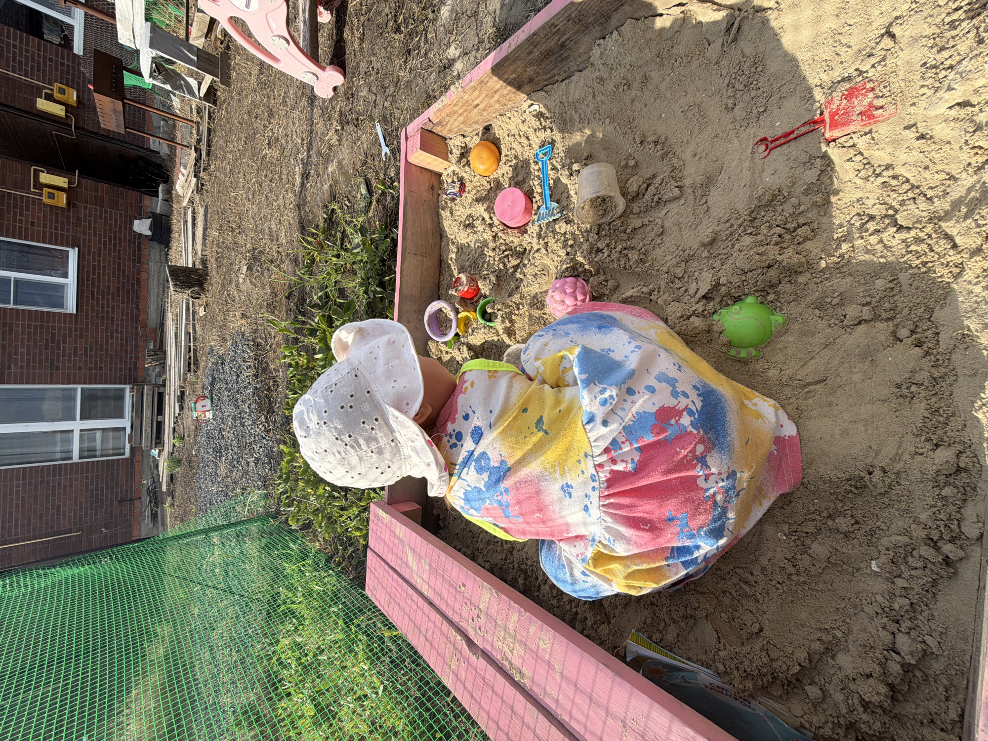 A child, wearing a white eyelet hat and a colorful, paint-splattered dress, lies on their side in a wooden sandbox filled with sand. Toys are scattered around the child and in the sand, including a blue plastic shovel, a red plastic shovel, a pink cup, an orange ball, and a green frog toy. The sandbox has pink wooden edges. To the left of the sandbox, a brick building with windows is visible, along with some green fencing and dry grass. A pink plastic slide is also partially visible in the background. The lighting suggests it is daytime, possibly late afternoon due to the shadows. The scene appears to be an outdoor play area.