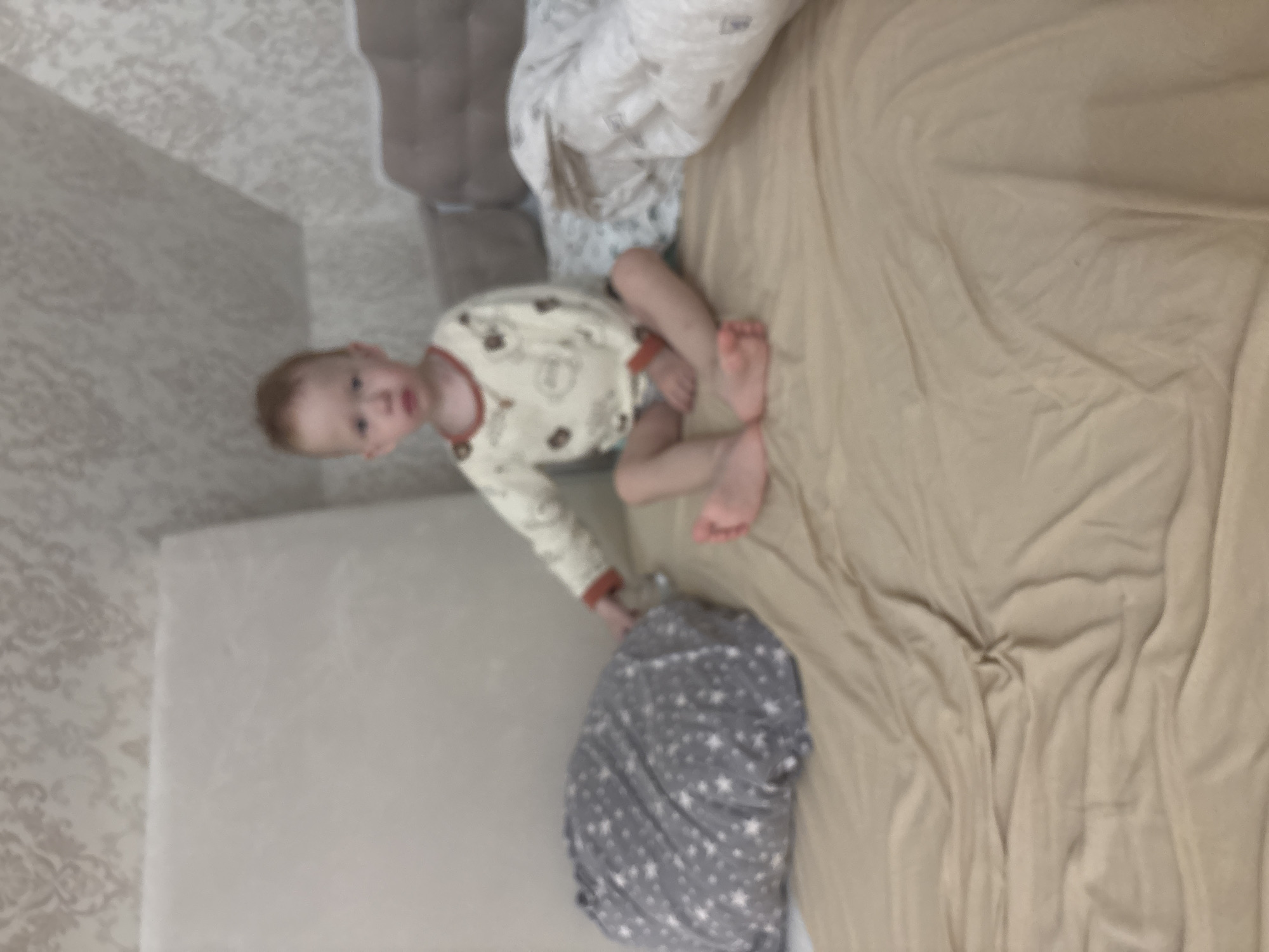 A child is on a bed. The child is wearing a white onesie with animal designs and has red hair. The bed is covered with a tan-colored sheet and there is a pillow with a white and gray patterned cover. The bed is next to a cushioned bench. The background includes a wall with patterned wallpaper. The location is Ферма, Russia.
