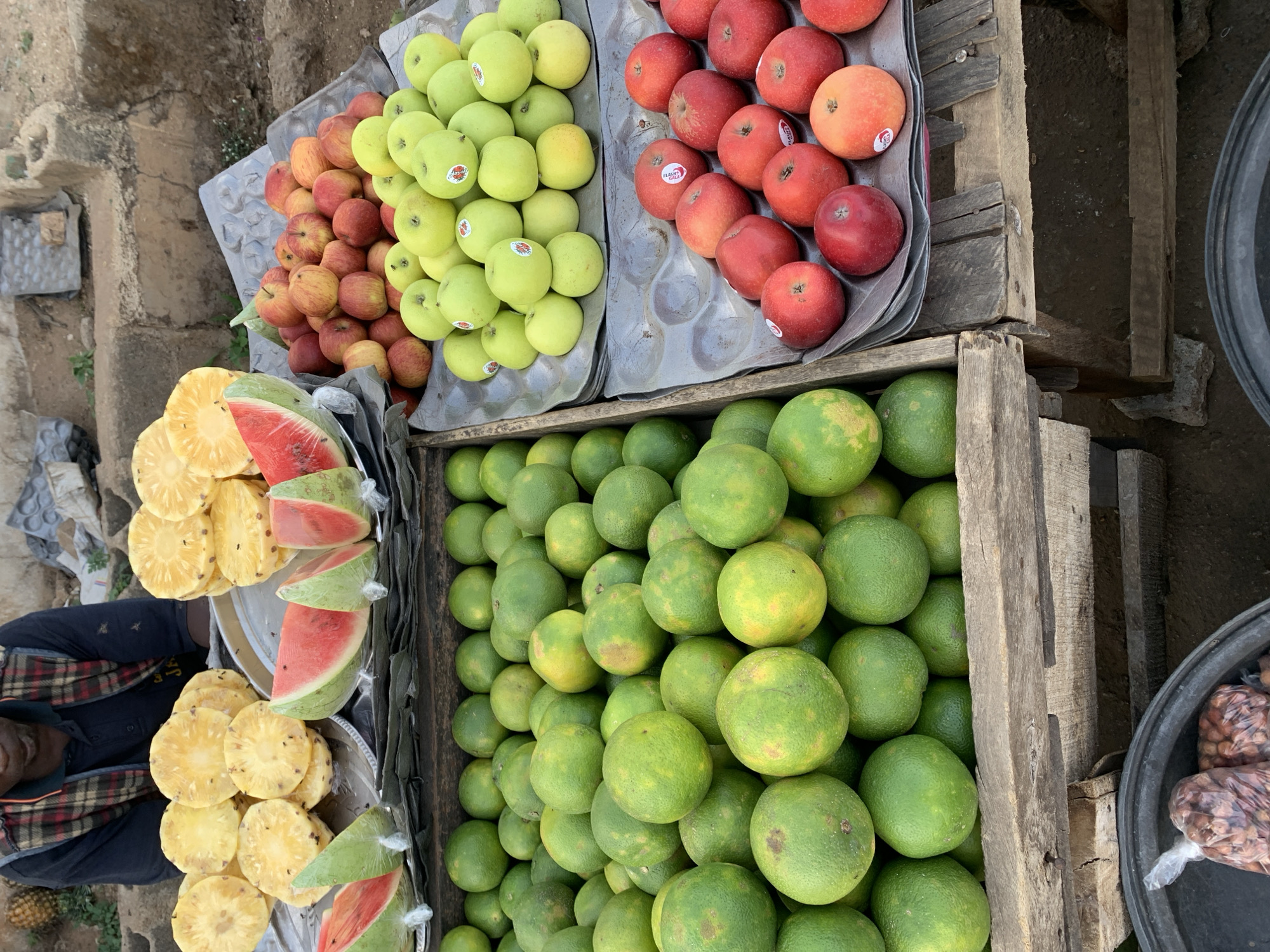A street vendor in New Stadium, Nigeria, displays a variety of fresh fruits. Crates and trays hold neatly arranged apples (red and green), whole green citrus fruits, sliced pineapples, and wedges of watermelon wrapped in plastic. A partial view of the vendor shows them seated, wearing a dark shirt and plaid jacket. To the side, a black tub contains bags of nuts. The background reveals a weathered, urban environment with visible brick and concrete.