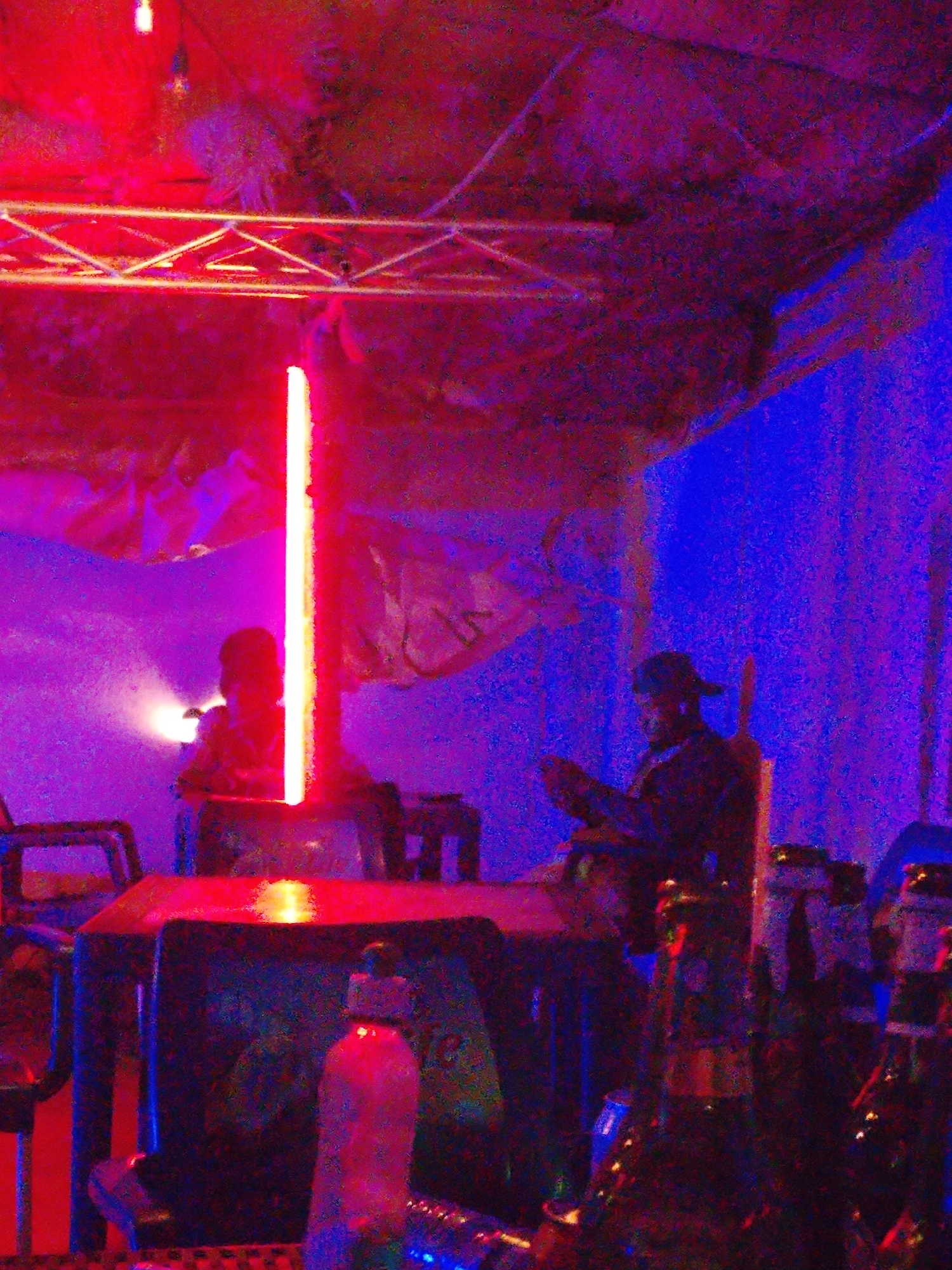 A dimly lit interior scene is captured, predominantly in red and blue hues. On the right, a figure wearing a cap is seated, possibly holding a device. In the center, a glowing vertical light fixture dominates the view, behind which another person is visible. A table is in the foreground, littered with bottles and containers. Overhead, a metal structure is visible. The location in the city is Unknown, Unknown.