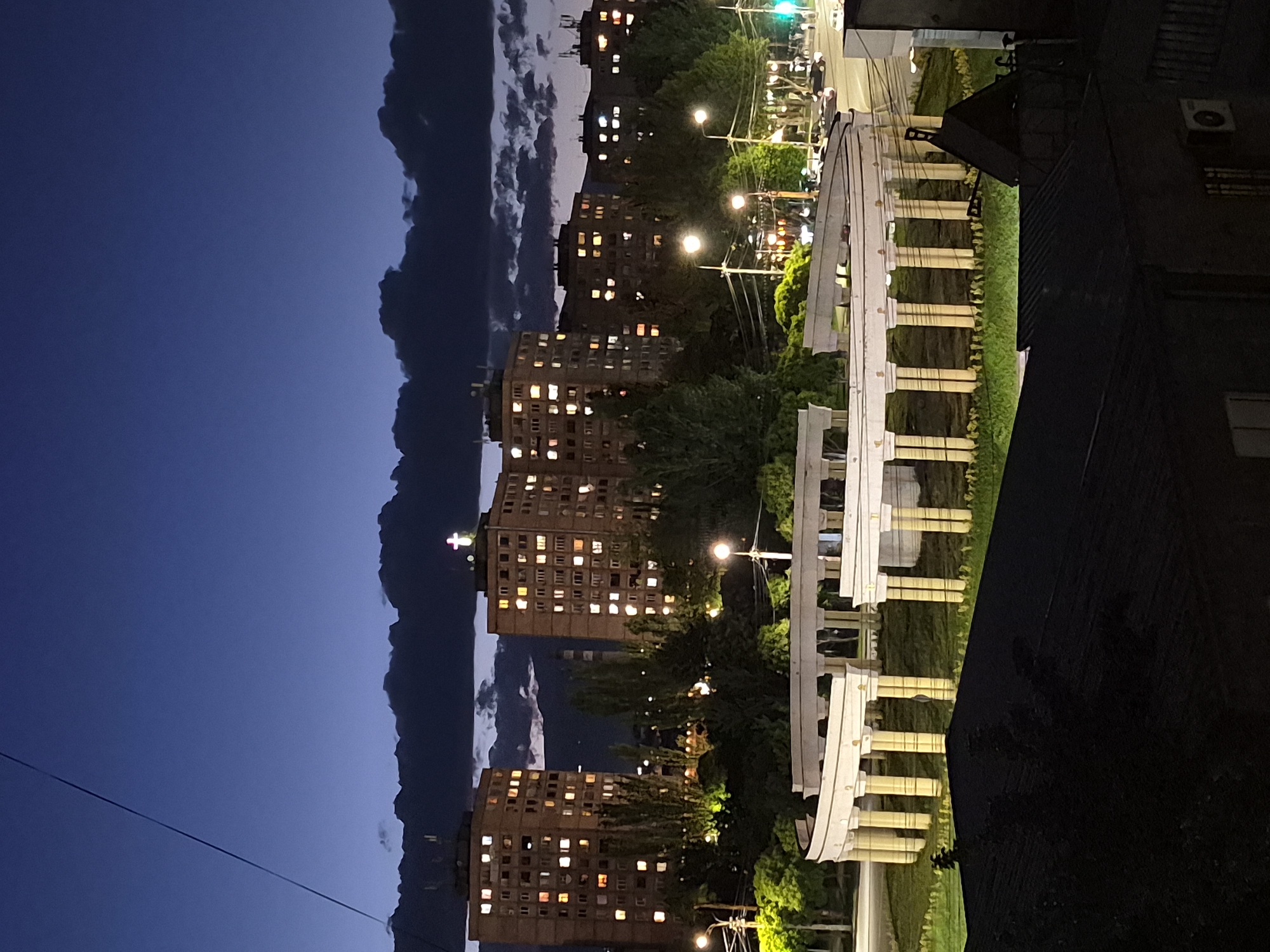 This image captures an urban scene at twilight or night, set in Yerevan, Armenia. The primary subjects are several multi-story residential buildings, a distinctive archway structure, and lush greenery, all under a darkening sky.
On the left side of the frame, multiple large apartment buildings, likely constructed from a dark-colored material like brick, rise into the sky. Many of their windows are illuminated, suggesting activity within the residences. A faint, cross-shaped light can be seen on top of a distant building or structure. The sky above is a deep blue, indicative of early night, with dark, fluffy clouds clustered along the upper left. A thin power line crosses the upper left portion of the sky.
In the mid-ground, extending towards the right, is a prominent white structure resembling a semi-circular or elliptical archway supported by pillars. This architectural feature appears to be part of a park or public space, surrounded by neatly maintained green lawns and numerous trees with vibrant green foliage. Several streetlights are scattered throughout the scene, casting warm glows on the trees and pathways. In the distance, a green traffic light is faintly visible.
The foreground on the bottom right is dominated by the dark, partially visible roof and side of a building, on which an air conditioning unit is mounted. The overall atmosphere is one of quiet urban life as evening descends, with artificial lights beginning to dominate the visual landscape. No visible text is discernible in the image.