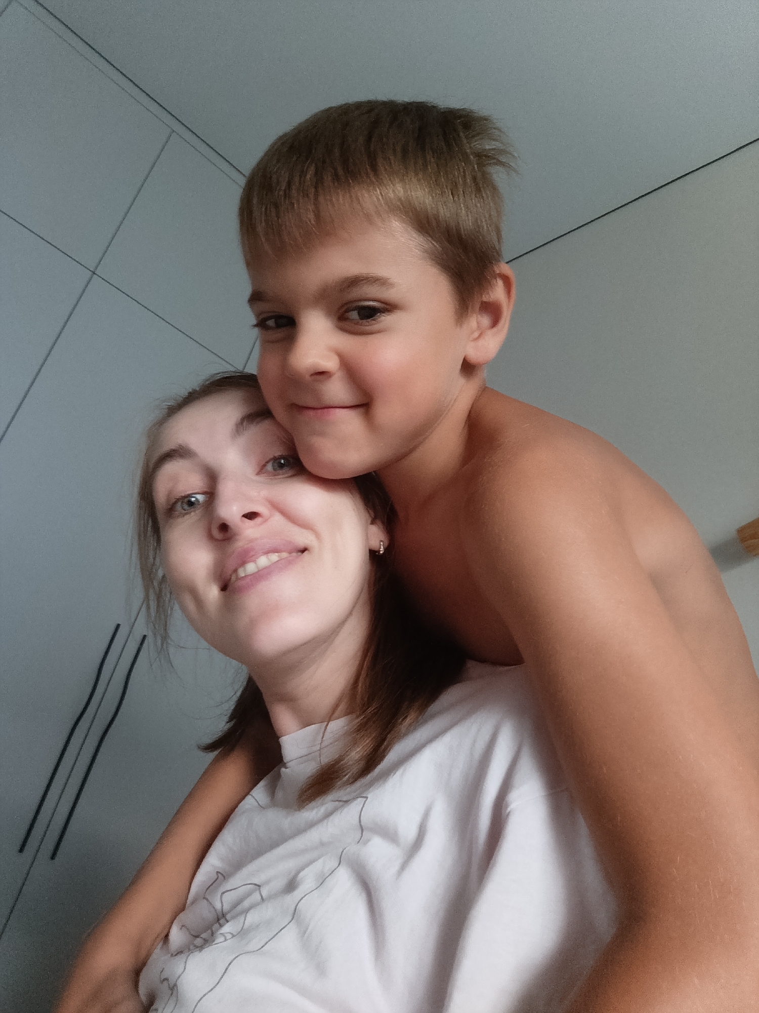 The image shows a close-up, selfie-style shot of a woman and a young boy, possibly mother and son, in an indoor setting. The boy is behind the woman, with his arms wrapped around her shoulders and his face resting on her head. Both individuals are smiling and looking towards the camera, conveying a sense of joy and affection. The woman has light brown hair and is wearing a white t-shirt with a faint line drawing on it. The boy has short brown hair and is shirtless.
The background appears to be a wall with a subtle grid pattern, possibly a closet door or a tiled surface, painted in a light grey hue. The lighting suggests daytime, though no specific time or weather conditions can be determined. The overall scene is intimate and personal, highlighting a close bond between the two subjects. There is no visible text in the image. The location context provided is Chaikovsky, Russia.