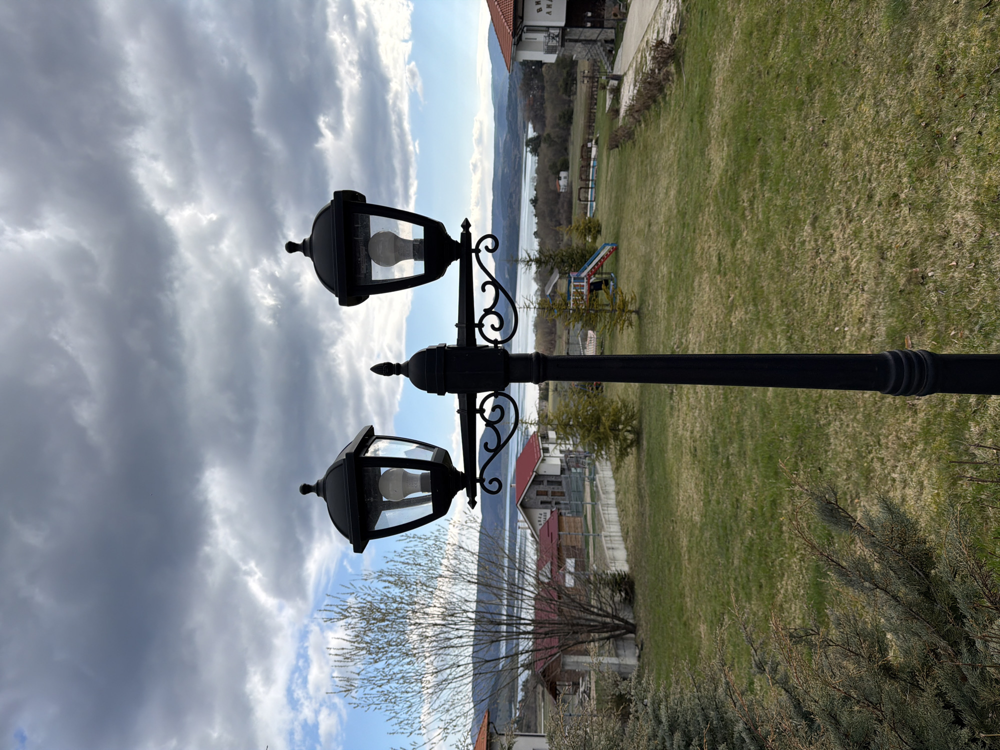 The image shows a black, three-lamp street light pole standing in a grassy area. The background includes a view of a lake or body of water and hills or mountains in the distance. Some buildings and structures are visible along the lakefront. The sky is cloudy. The overall setting appears to be in a residential area near a body of water. The location is described as Unknown, Unknown.