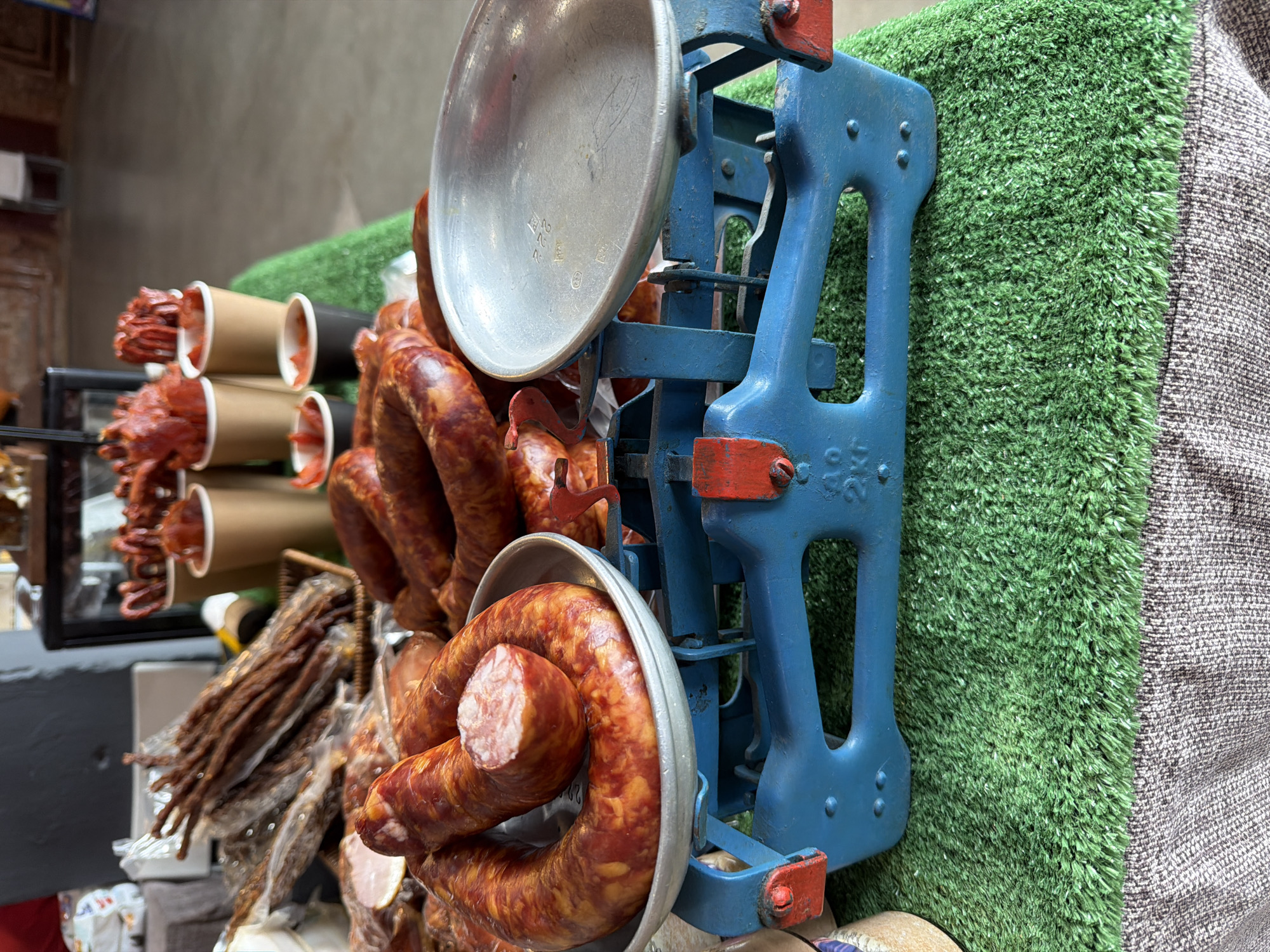 The image depicts a butcher shop display in what appears to be Saint Petersburg, Russia. A vintage, blue and red scale sits prominently, holding rings of sausage in its silver pans. Behind the scale, a wall covered with artificial green turf is seen next to grey fabric. Cups filled with what appears to be meat or sausages are lined up behind the scale, suggesting a variety of offerings. Wrapped sticks of meat are on display in the lower left corner, indicating a selection of dried meats. Overall, the scene suggests a close-up view of a curated presentation of meats for sale in an urban setting.