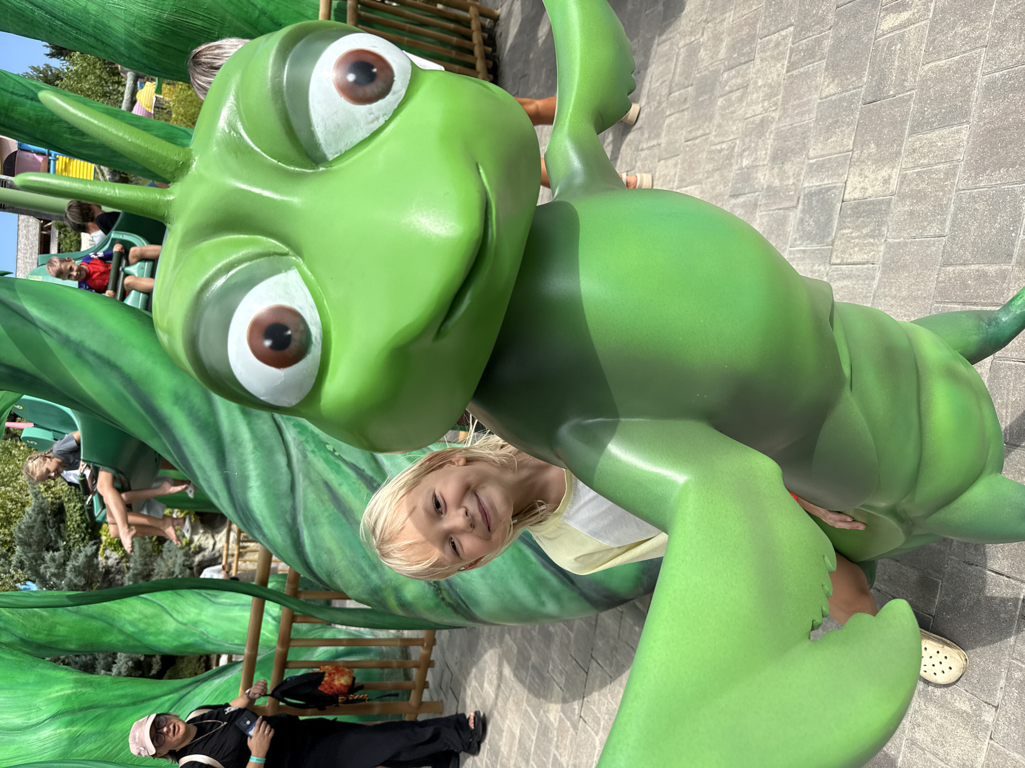 In this outdoor, sunny photograph, a young blonde child with a bright smile poses next to a large, cartoonish green insect statue. The child, wearing a yellow and white t-shirt, is positioned behind the statue's right leg, with their head and shoulders visible. The statue, a bright green, stylized insect with large, brown-eyed cartoon eyes, dominates the foreground. Its body is smooth and shiny, and it has large, claw-like appendages. In the background, a woman in a pink hat and sunglasses stands to the left, holding a phone and a bag. Further back, other children are visible on a green ride structure. The ground is paved with interlocking grey stones, and a wooden fence runs horizontally across the mid-ground. The environment suggests a theme park or amusement area with lush green foliage and vibrant structures. The location is not specified to be in the city of Симеизский поселковый совет, Ukraine, as no such location is depicted.