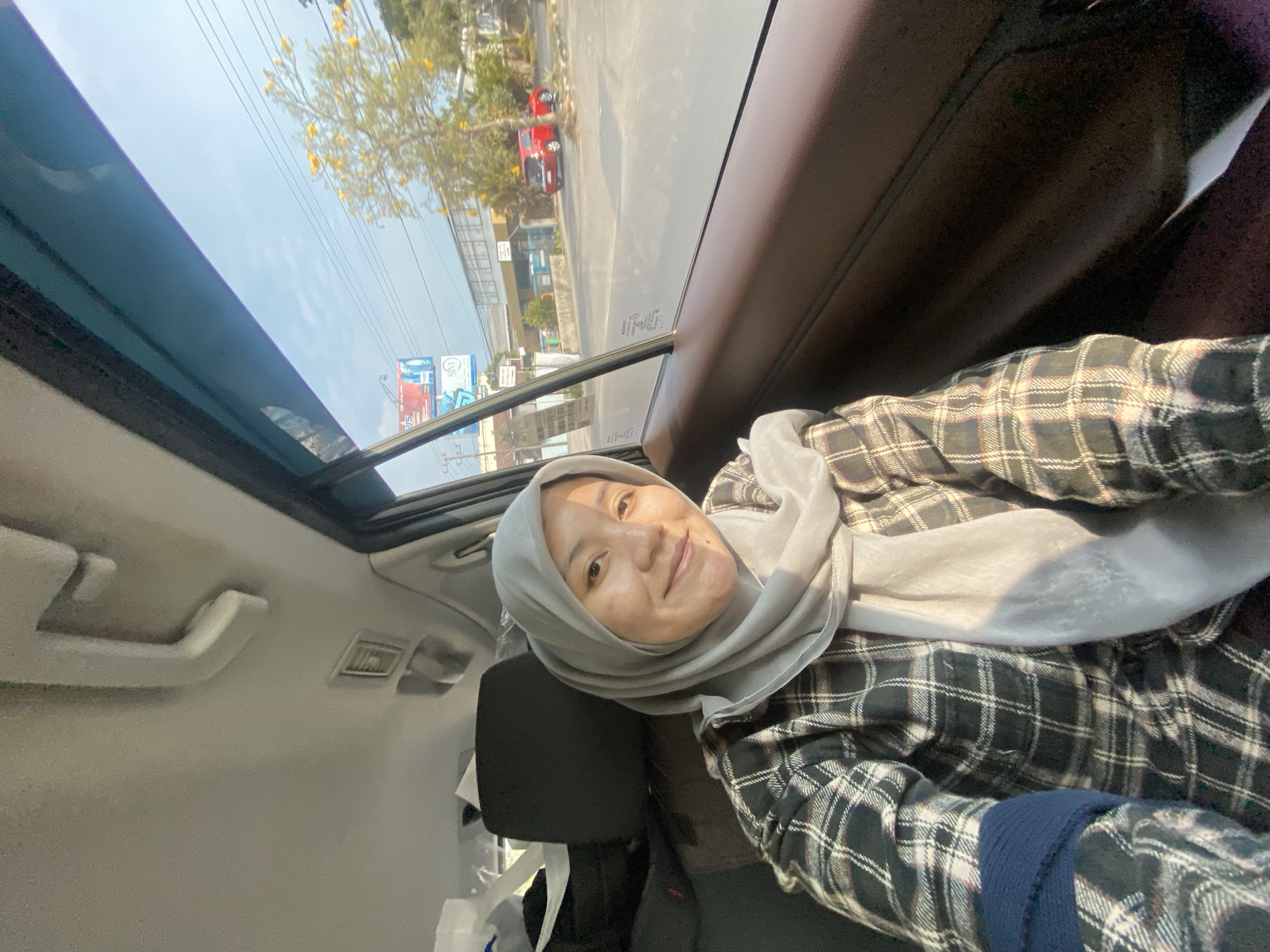 A woman wearing a light grey hijab and a plaid shirt is seated in the passenger seat of a car. She is looking at the camera and smiling. The interior of the car is visible, including the brown leather seat and the grey headliner. Through the window, a street scene in Mandar Sari, Indonesia, is visible. There are trees, buildings, power lines, and other cars on the road. The sun is shining, casting shadows on the scene.